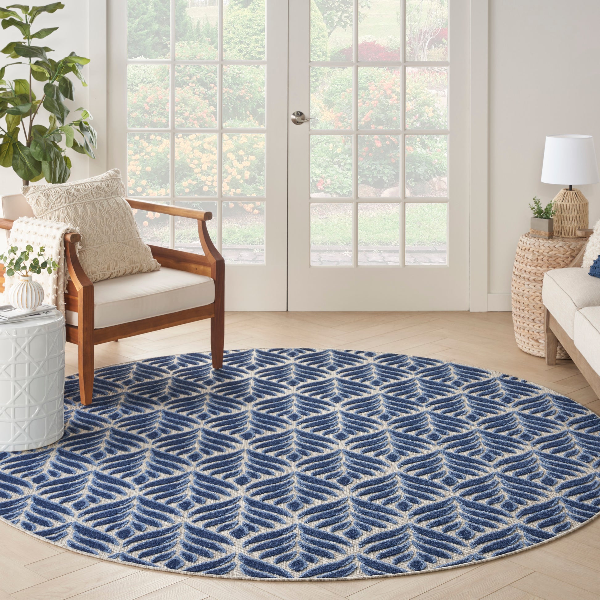 Round blue and white geometric rug on light wood floor in bright living room with wooden armchair, plants, and French doors.