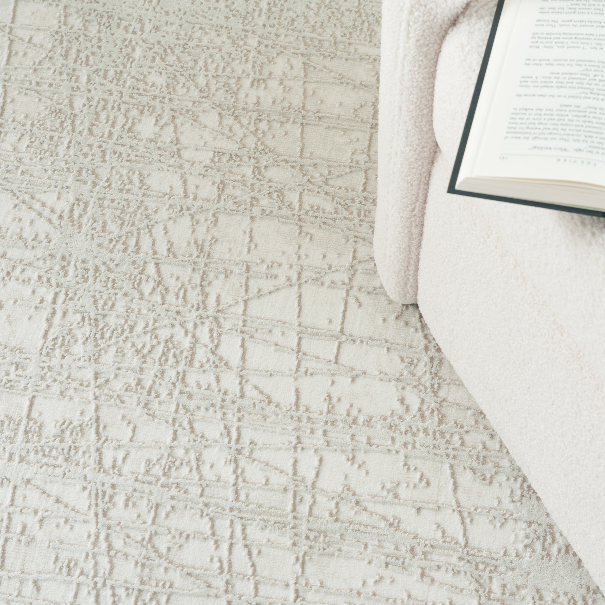 Soft beige textured area rug with a subtle geometric pattern, a cream throw and open book creating a cozy reading corner.