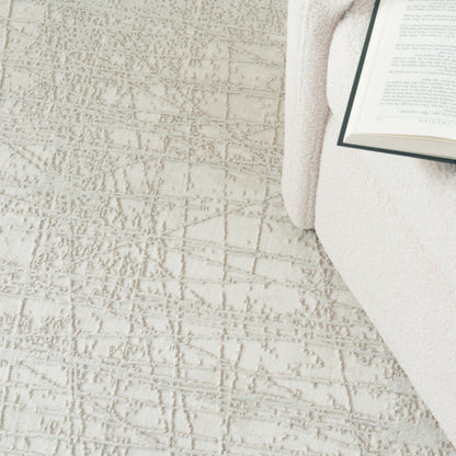 Soft beige textured area rug with a subtle geometric pattern, a cream throw and open book creating a cozy reading corner.