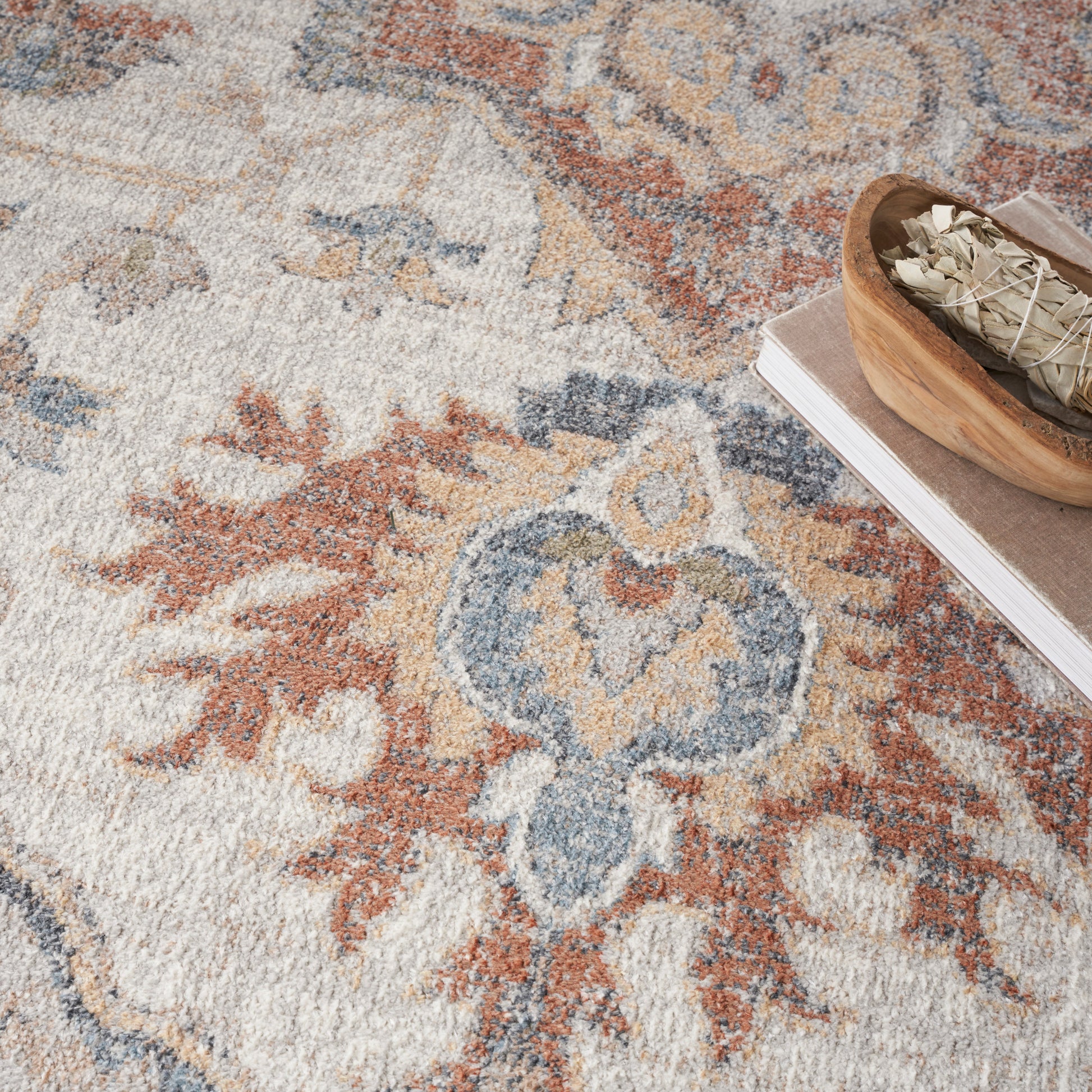 Persian-style area rug in cream, blue and rust with a floral medallion; wooden bowl holding sage bundles on a book.