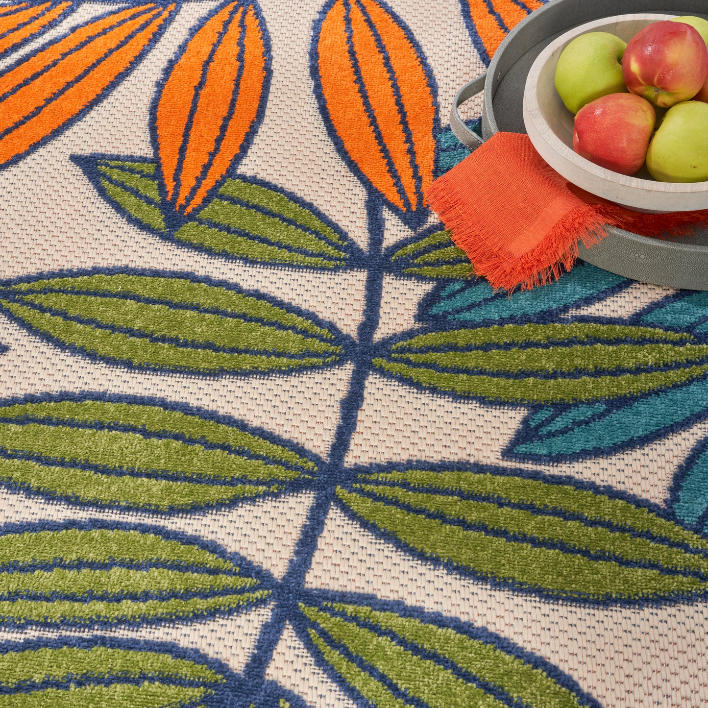 Close-up of a woven rug with orange floral shapes and green leaves; a metal fruit bowl with apples nearby.