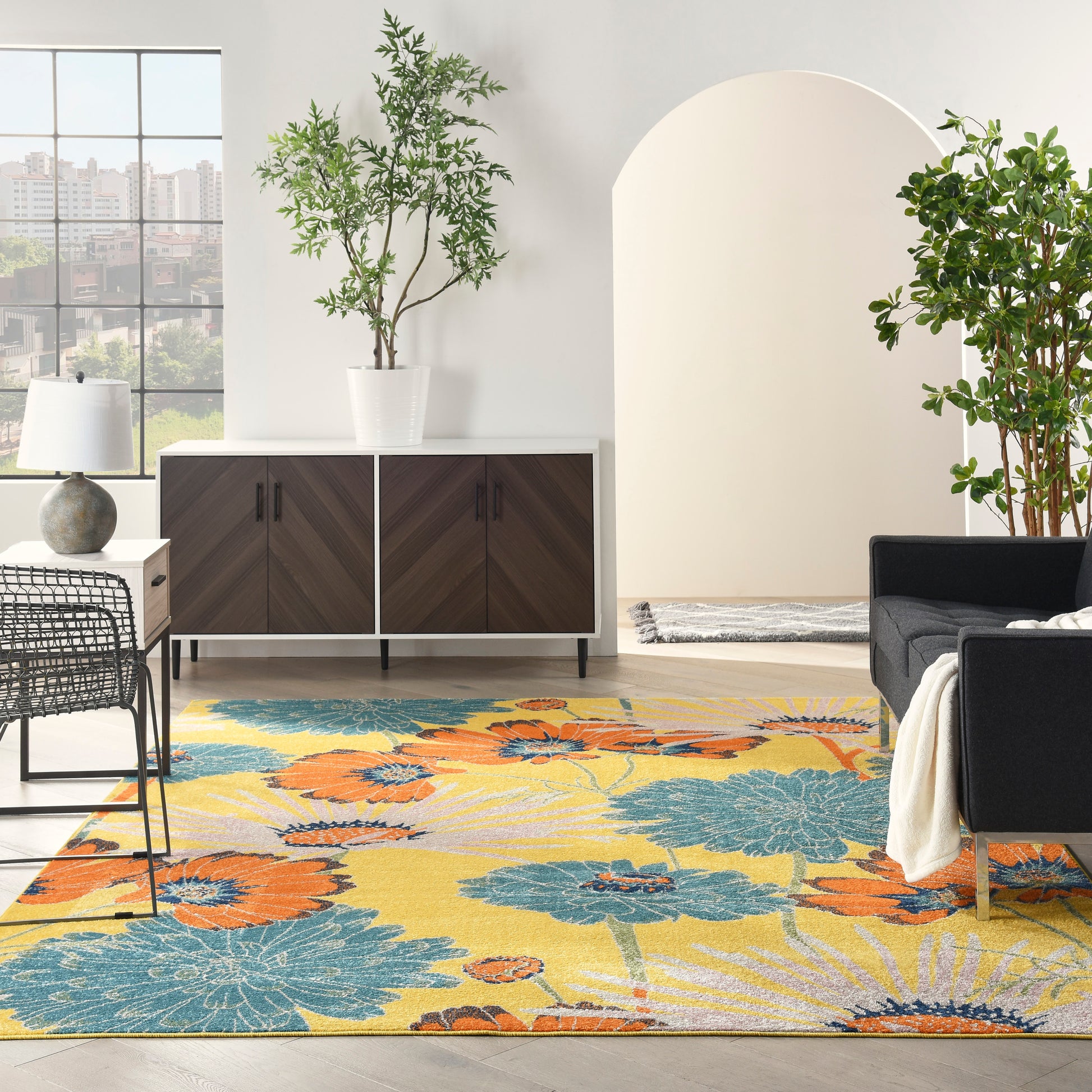 Bright modern living room with chevron-wood cabinet, colorful floral rug in yellow, orange and teal, black seating, and potted plants.