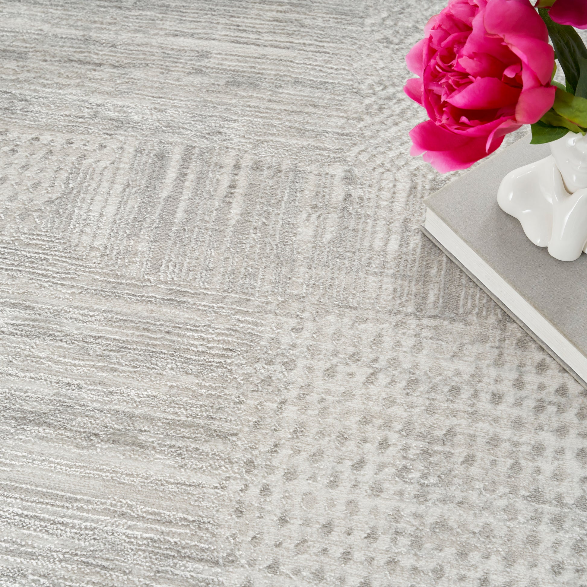 Pink rose beside a white ceramic sculpture on a gray hardcover book, atop a textured light-gray rug.