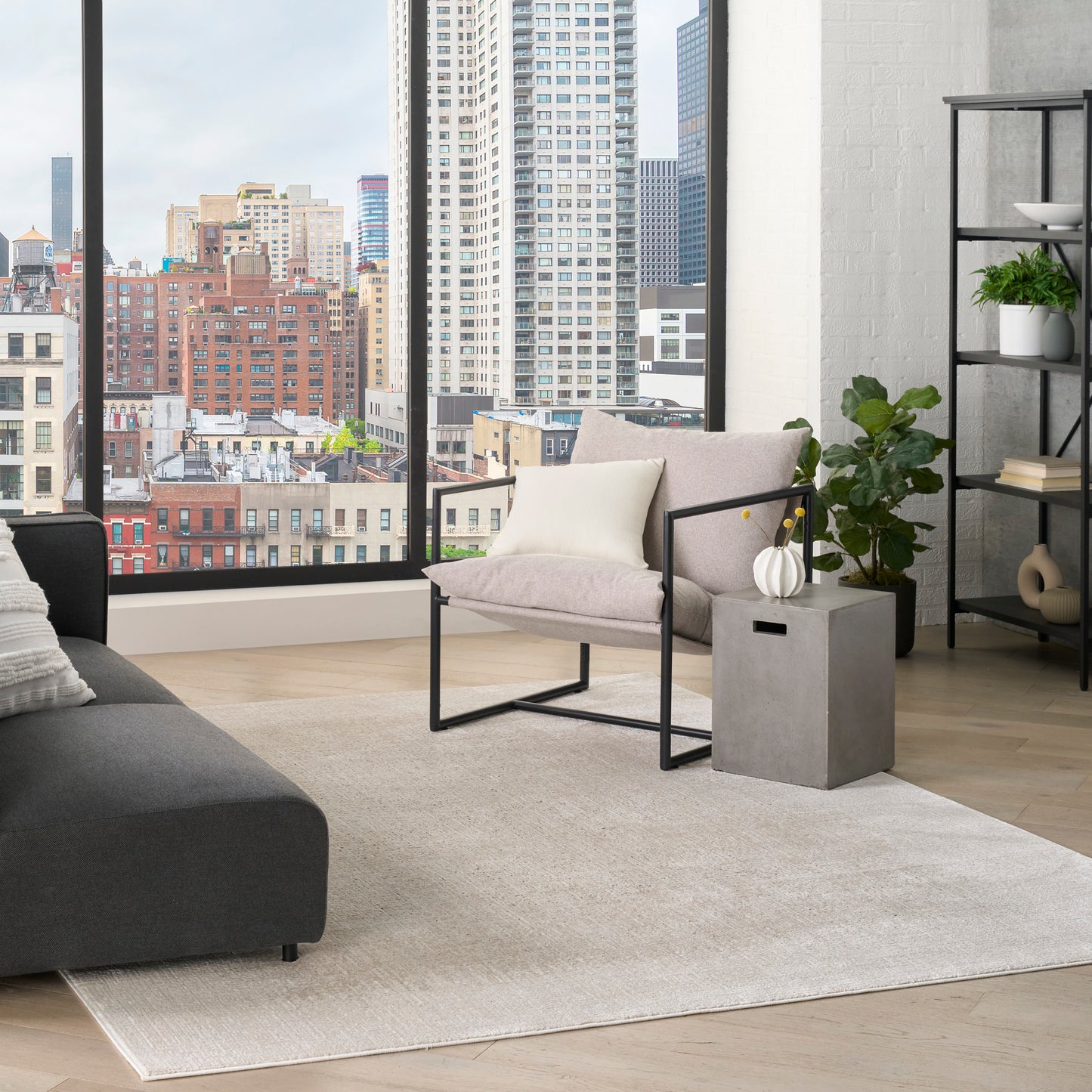 Modern 6' x 9' rug in a living room on the floor, accompanied by a gray sofa, beige metal-frame chair, a concrete table with a vase, and potted plants.