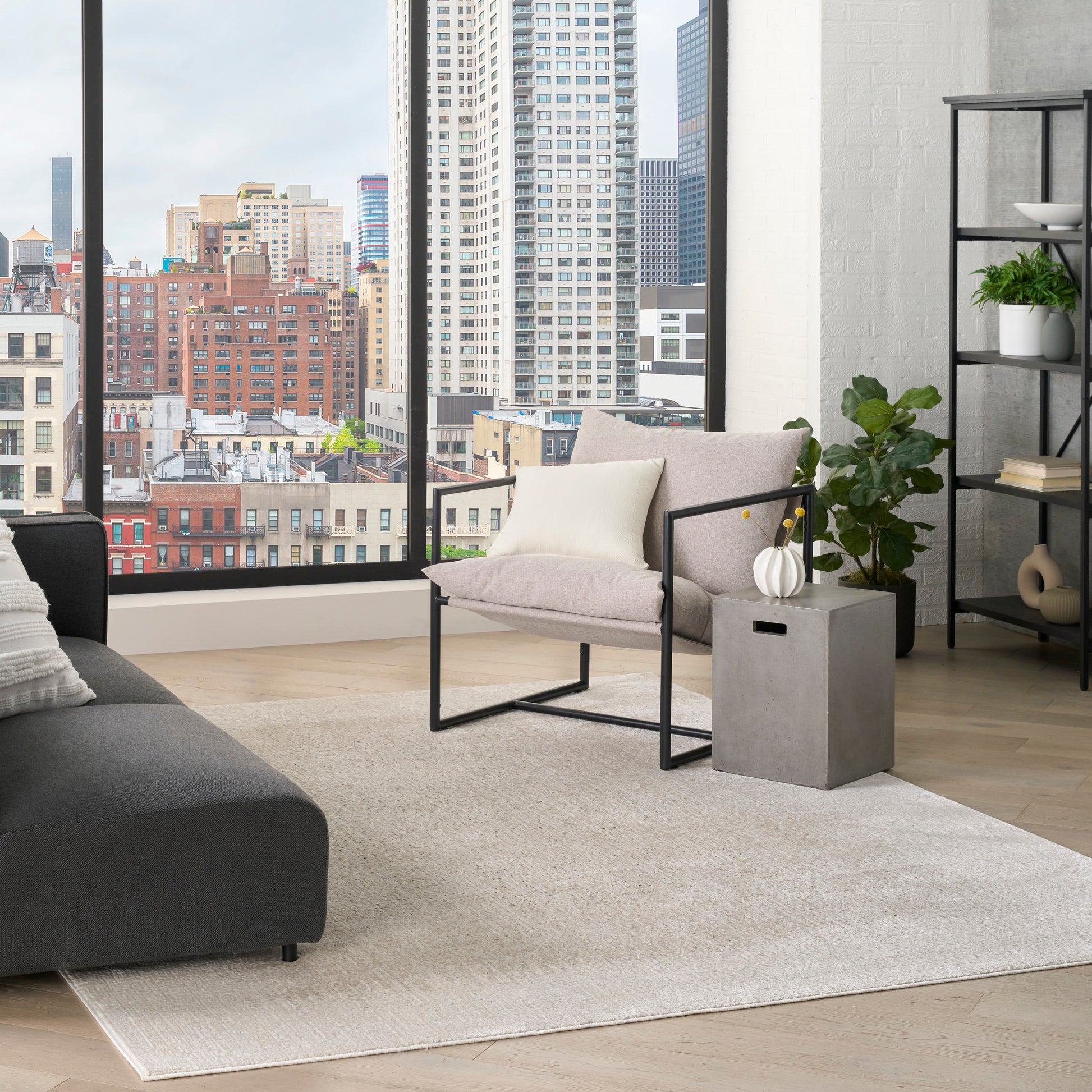 Modern 6' x 9' rug in a living room on the floor, accompanied by a gray sofa, beige metal-frame chair, a concrete table with a vase, and potted plants.