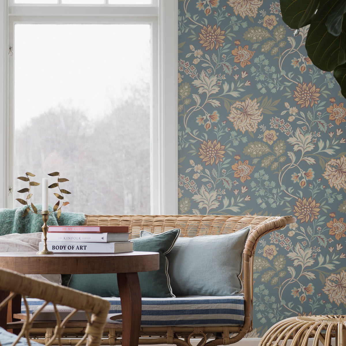 Wicker sofa with teal cushions in a sunlit room, blue floral wallpaper, wooden coffee table with books and a candle holder.