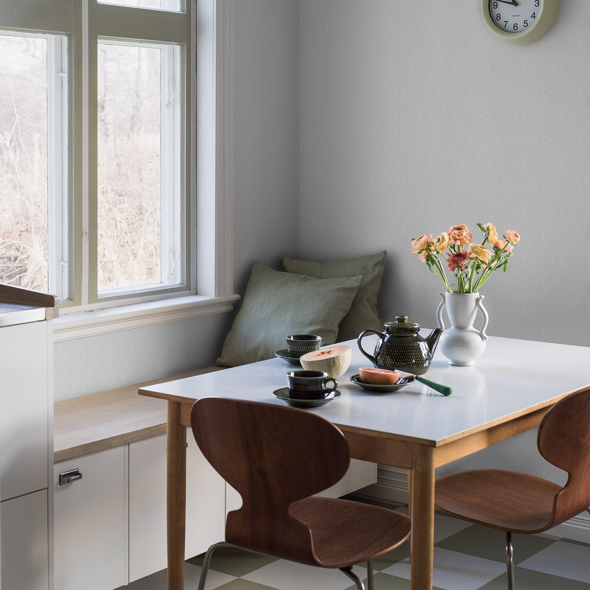 Bone-colored wallpaper behind a sunlit dining nook; white table, brown wooden chairs, teapot, cups and a vase of peach flowers.