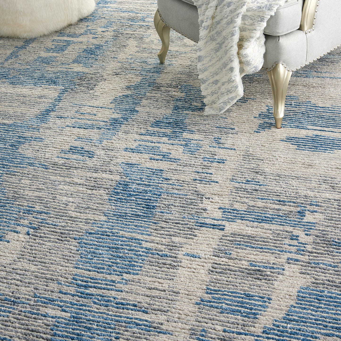 Soft beige and blue patterned area rug with abstract motifs, shown with a plush throw and ornate silver-legged sofa.