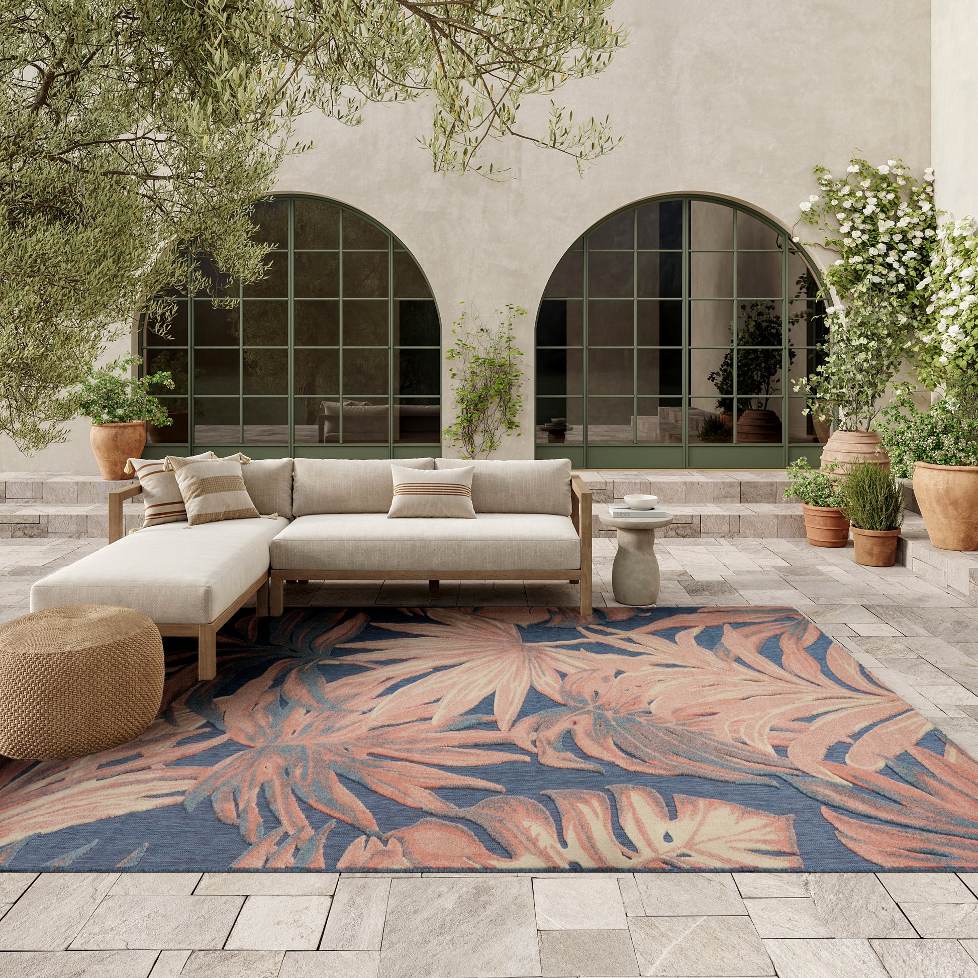 Outdoor lounge with beige fabric sectional on wood frame, tropical blue-pink leaf-pattern rug, arched windows, potted plants.