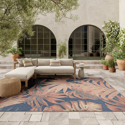 Outdoor lounge with beige fabric sectional on wood frame, tropical blue-pink leaf-pattern rug, arched windows, potted plants.