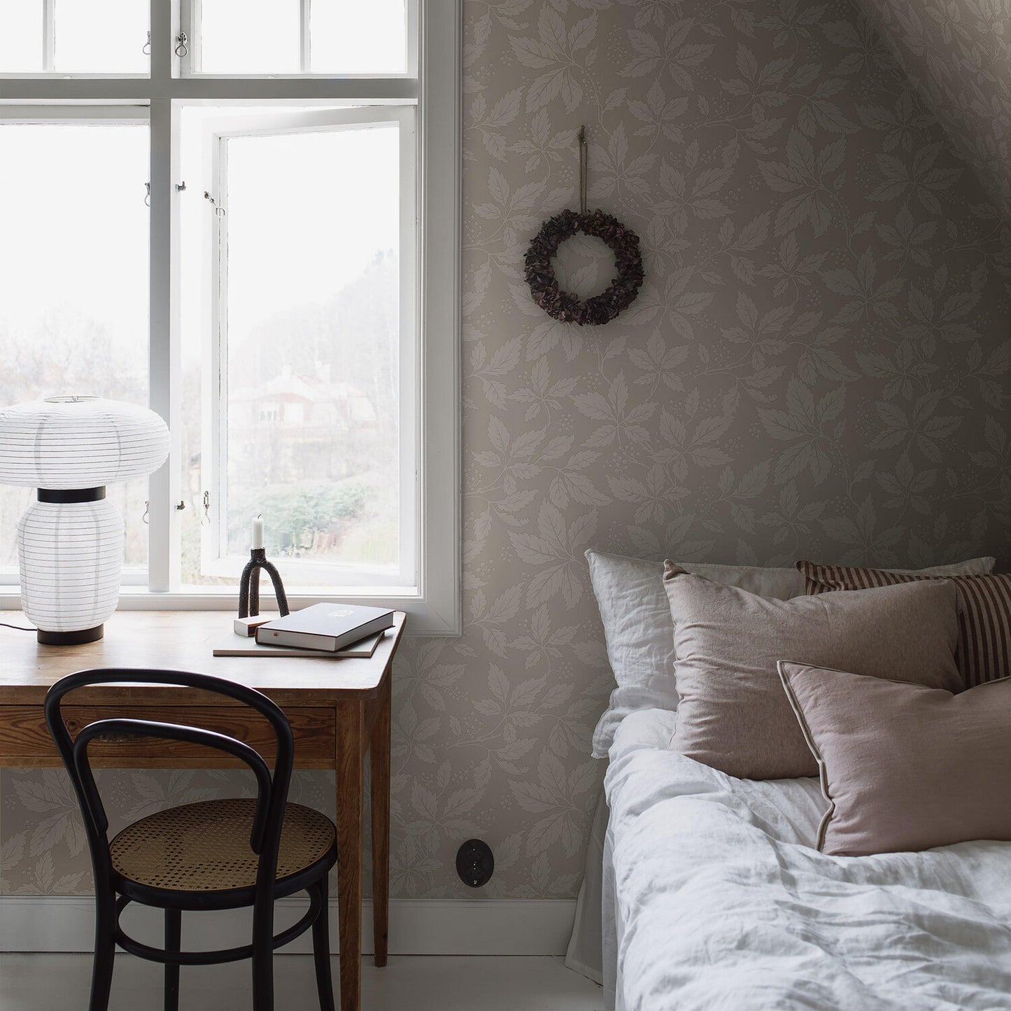 Cozy bedroom nook with wooden desk by a sunlit window, white lantern lamp, notebook, black chair, stone-pattern wallpaper, wreath, and bed with beige cushions.