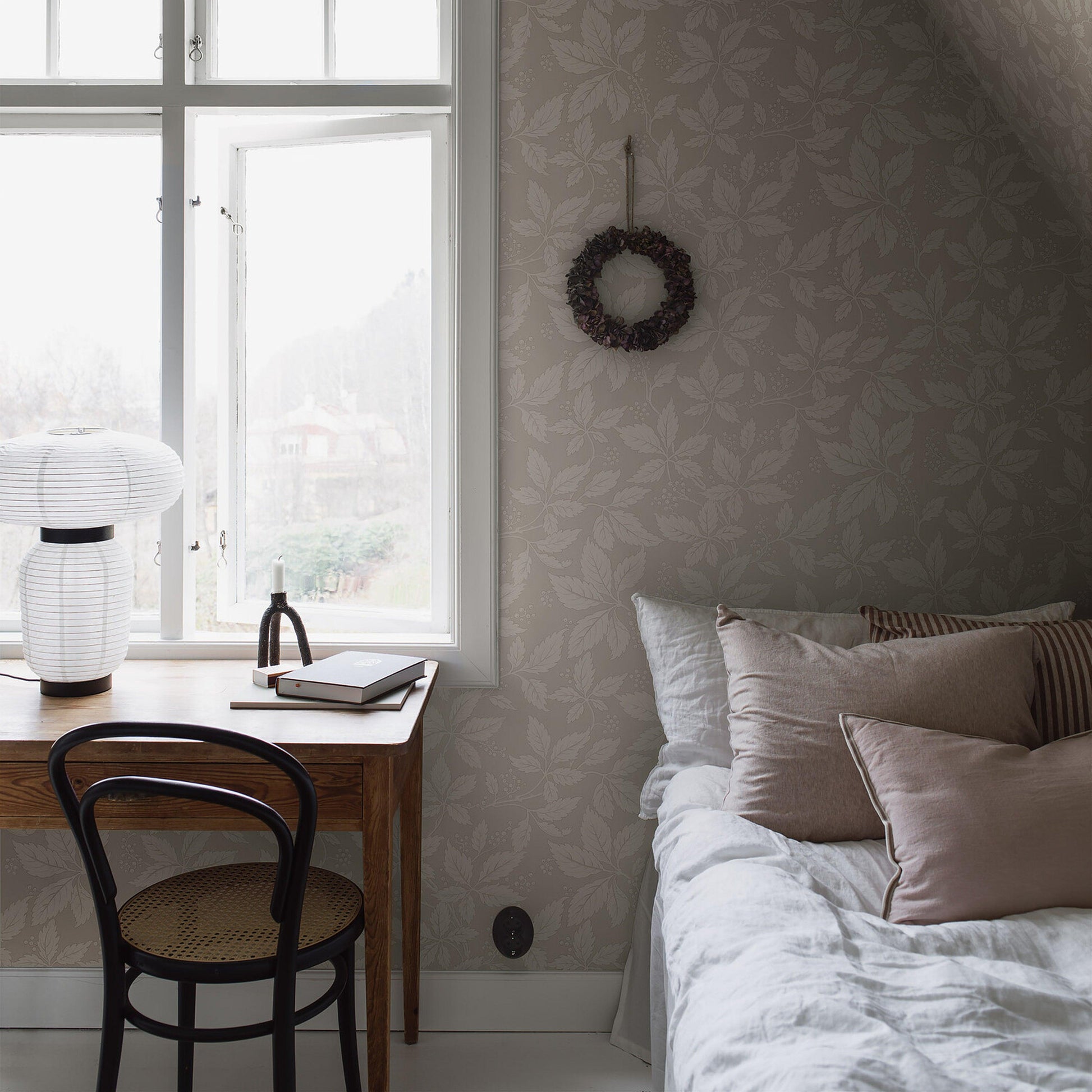 Cozy bedroom nook with wooden desk by a sunlit window, white lantern lamp, notebook, black chair, stone-pattern wallpaper, wreath, and bed with beige cushions.