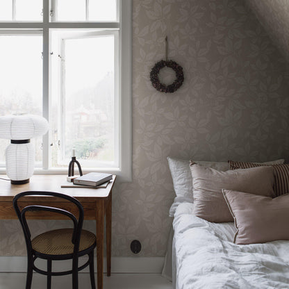 Cozy bedroom nook with wooden desk by a sunlit window, white lantern lamp, notebook, black chair, stone-pattern wallpaper, wreath, and bed with beige cushions.