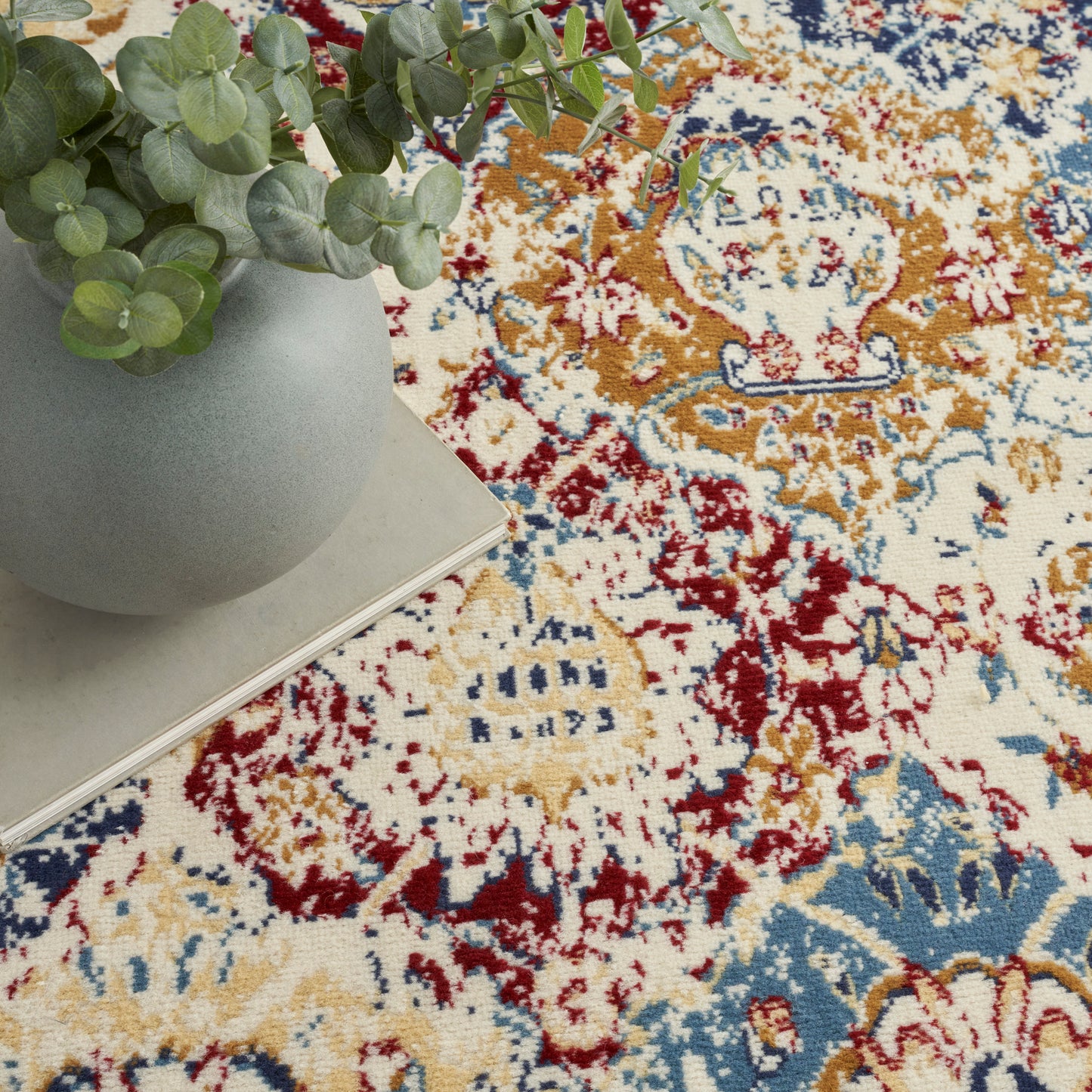Grey ceramic vase with eucalyptus stems on a stack of books over a vibrant oriental rug with gold, red and blue motifs.
