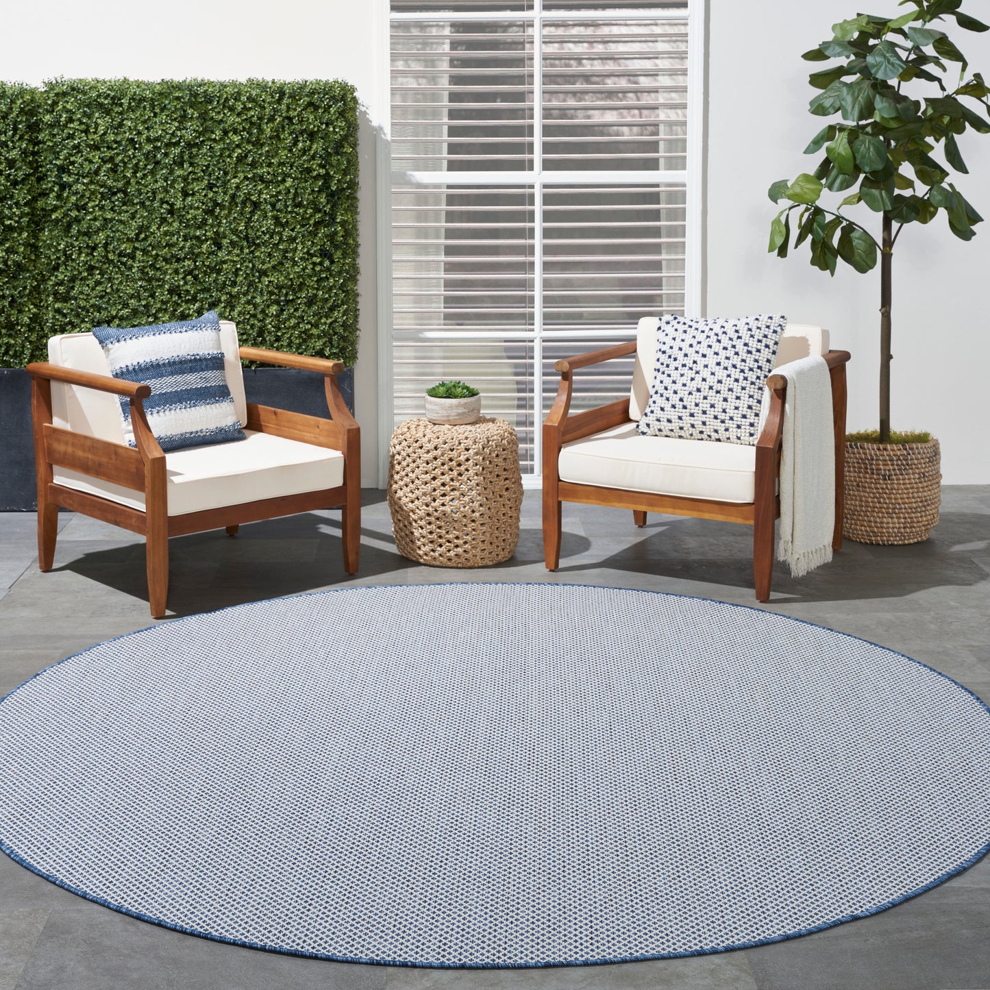 Outdoor patio with wooden-frame armchairs, white cushions, blue pillows, round blue rug, wicker side table and plants.
