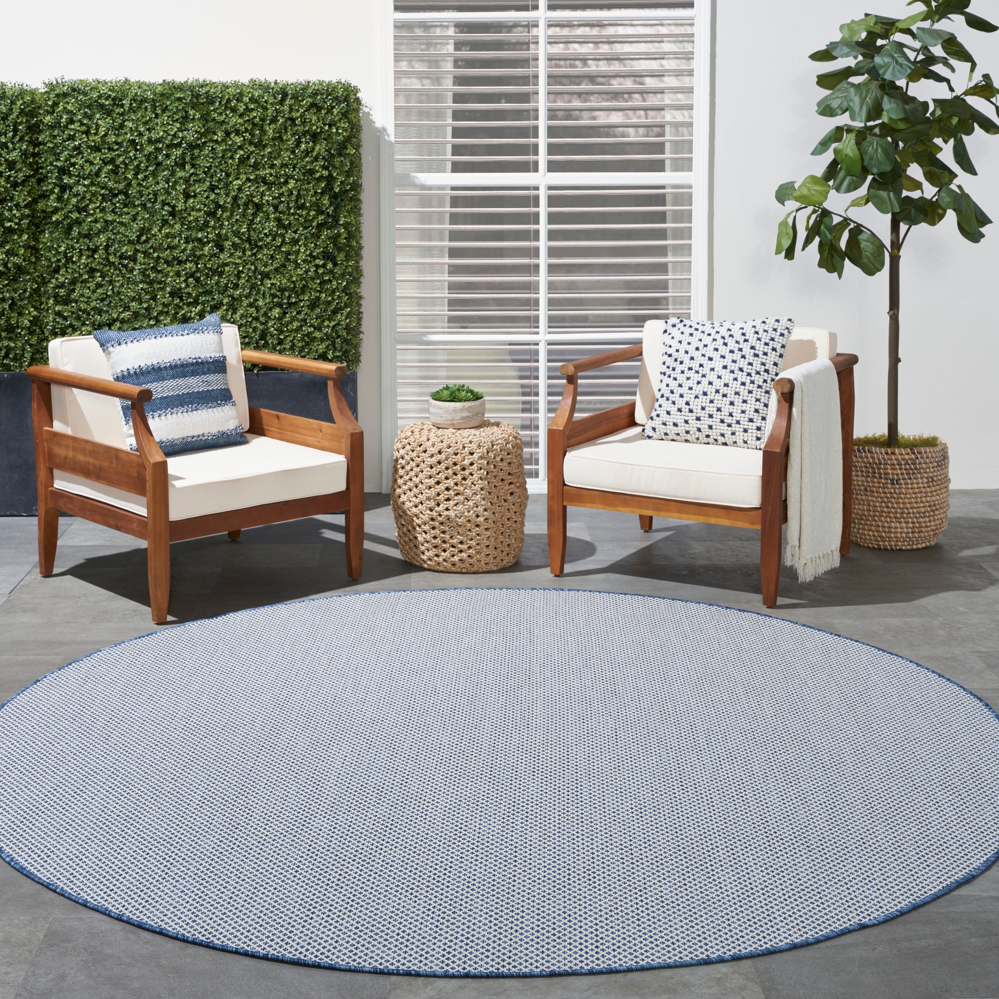 Outdoor patio with wooden-frame armchairs, white cushions, blue pillows, round blue rug, wicker side table and plants.