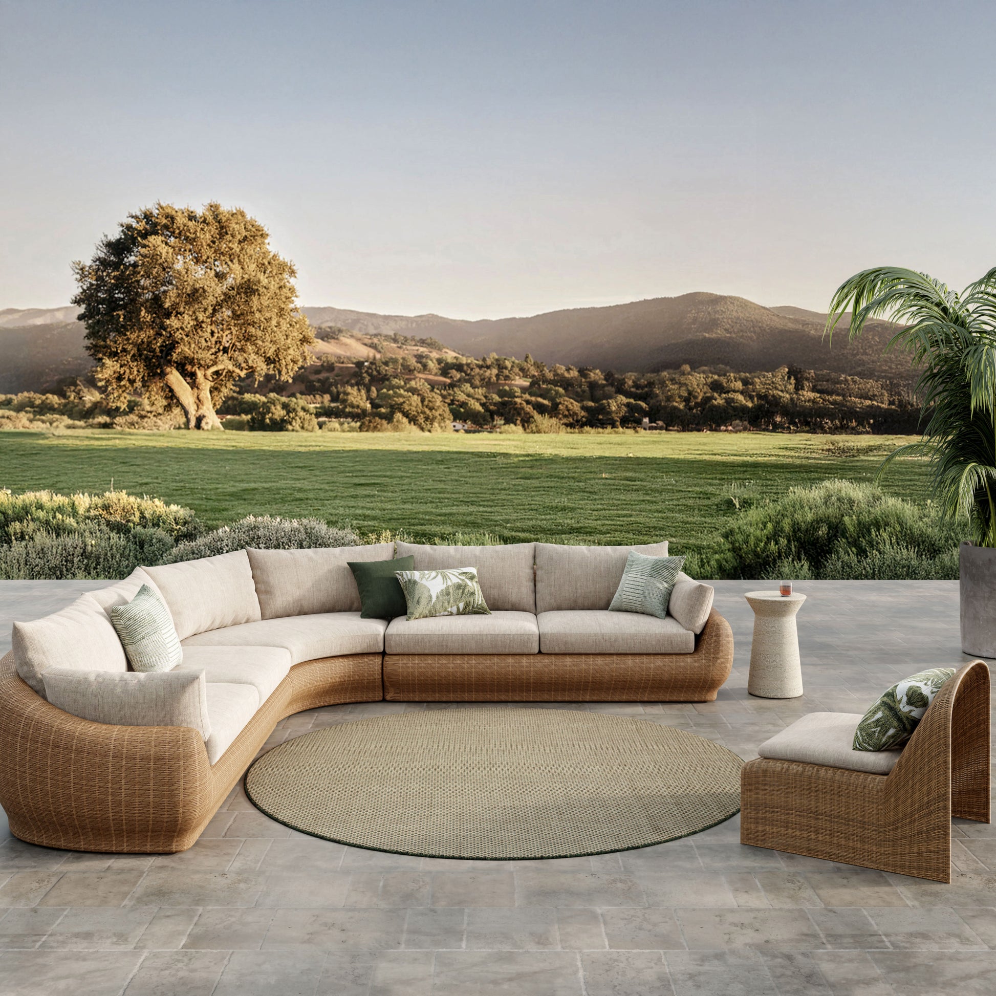Outdoor rattan sectional with cream cushions and green pillows on a circular beige rug, patio scene overlooking countryside.