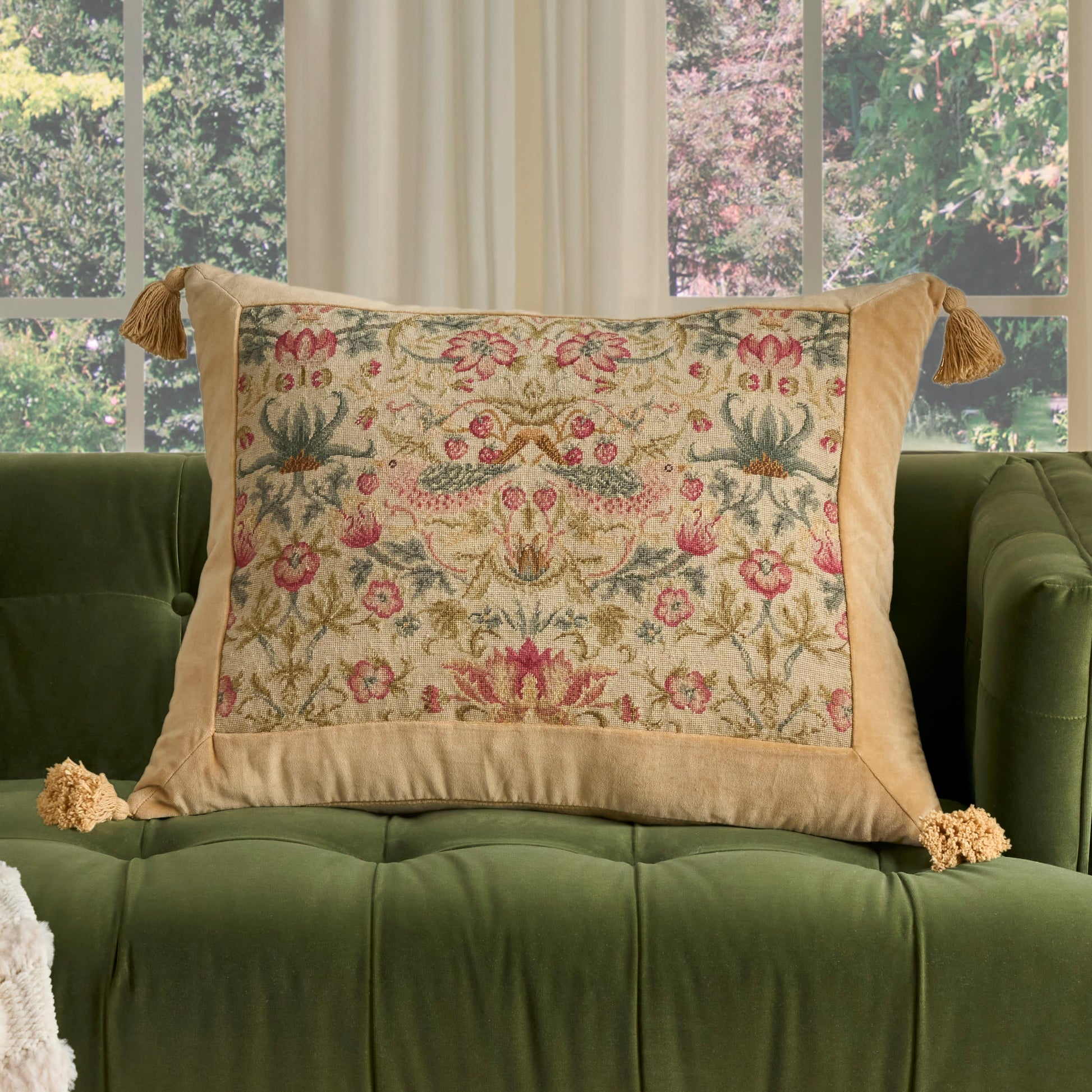 Beige embroidered floral tapestry pillow with pink and green flowers and tassels, resting on a green tufted sofa.