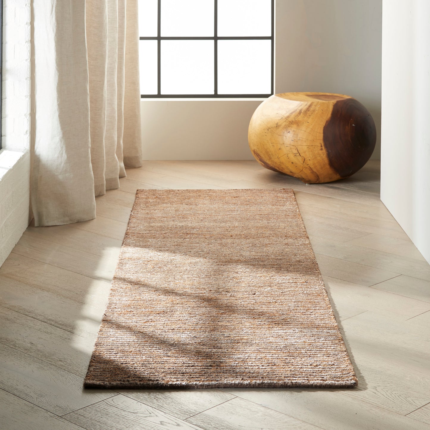 Natural jute rug runner on a light wood floor beside beige curtains and a round wooden stool by a grid-pane window.