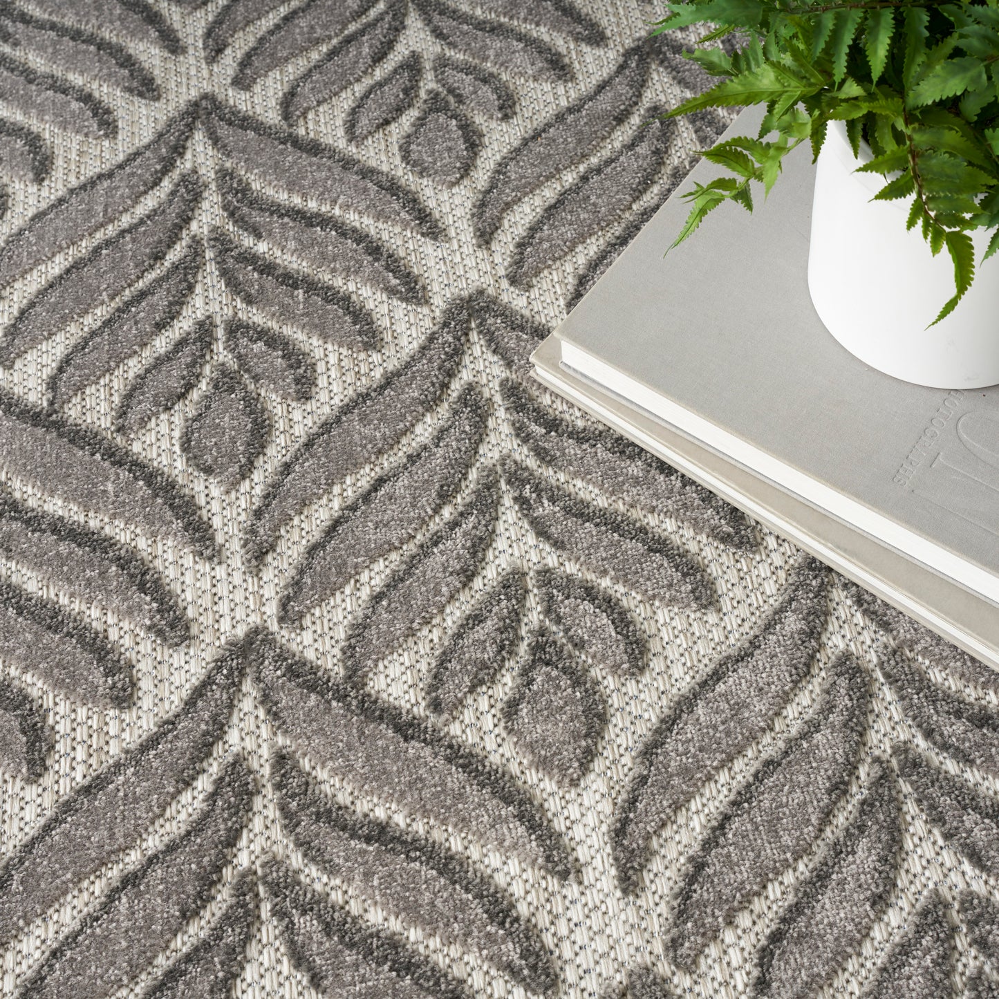 Gray leaf-patterned rug with textured pile; white planter with a green fern beside a book stack.