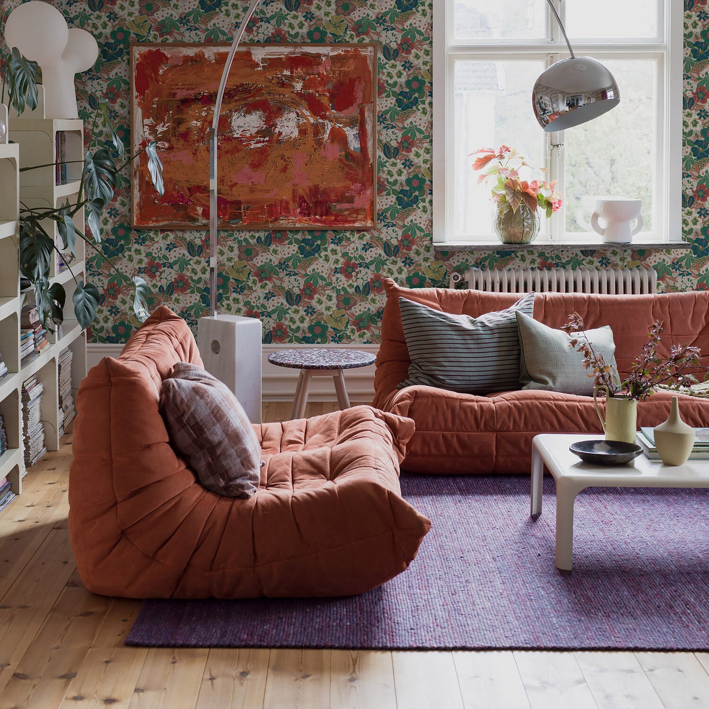 Cozy living room with orange tufted sofa & lounge chair on a purple rug, floral wallpaper & white coffee table.