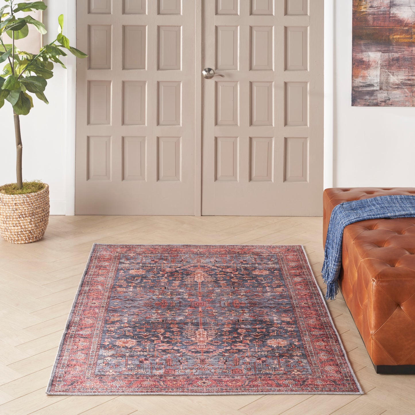 Oriental rug with red-blue pattern on light wood floor, beige paneled doors, potted plant, tufted brown leather sofa.
