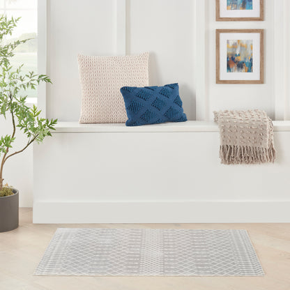White bench with beige knitted pillow, navy lattice pillow, fringed beige throw, plant, framed art, and gray geometric rug.