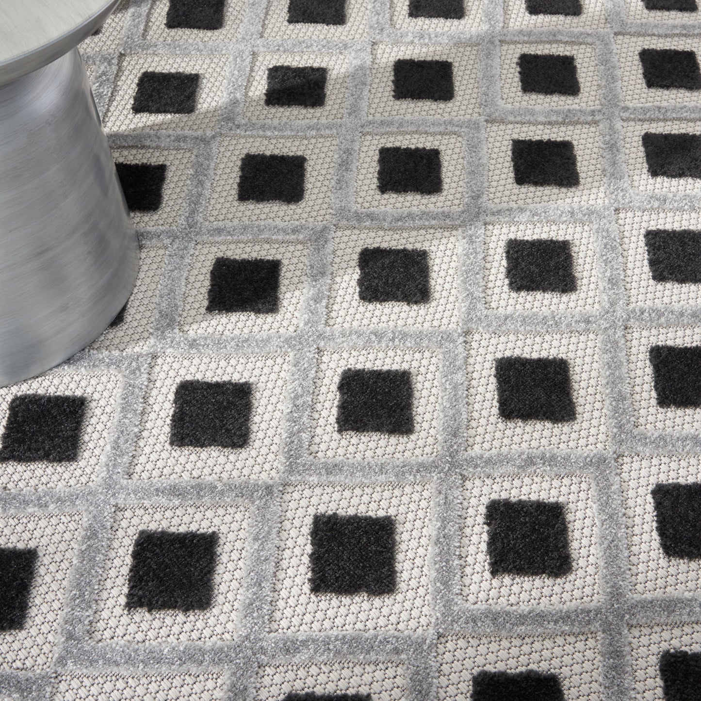 Geometric gray woven rug with repeating light-gray squares and black centers; modern floor mat, metal roll visible on left.