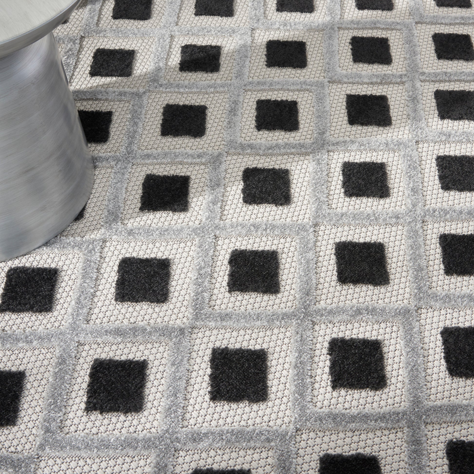 Geometric gray woven rug with repeating light-gray squares and black centers; modern floor mat, metal roll visible on left.