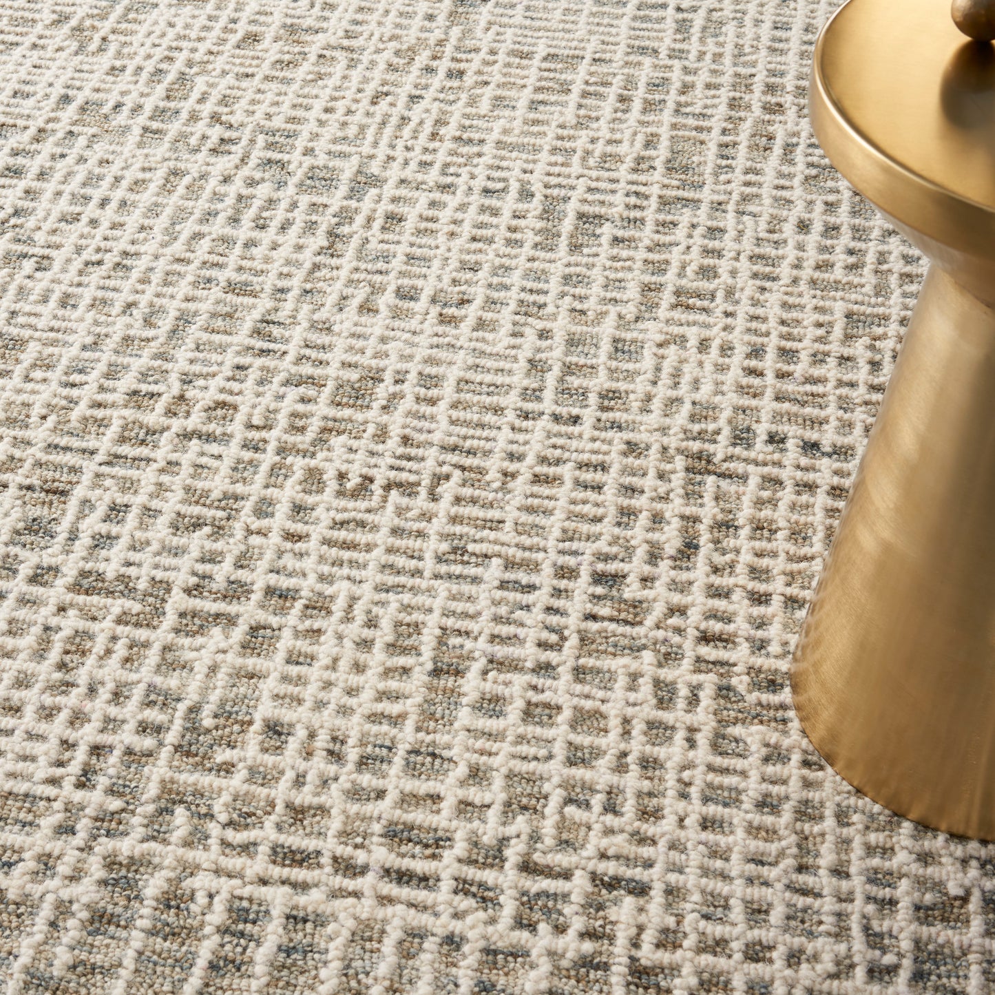 Beige patterned area rug with a gray geometric lattice design beside a gold metal table leg.