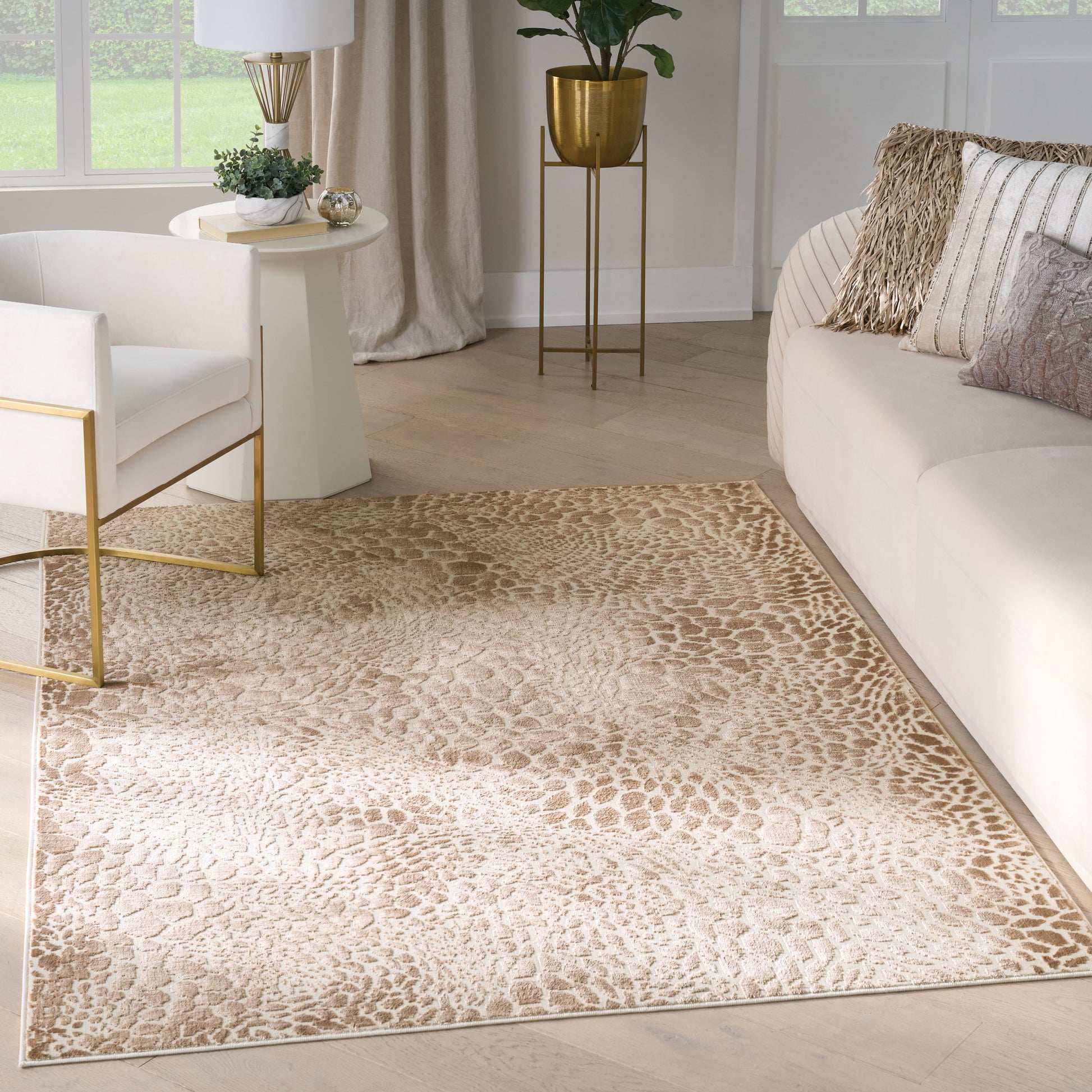 Neutral living room with beige patterned rug, ivory sofa, white chair with gold metal legs, and gold plant stand.