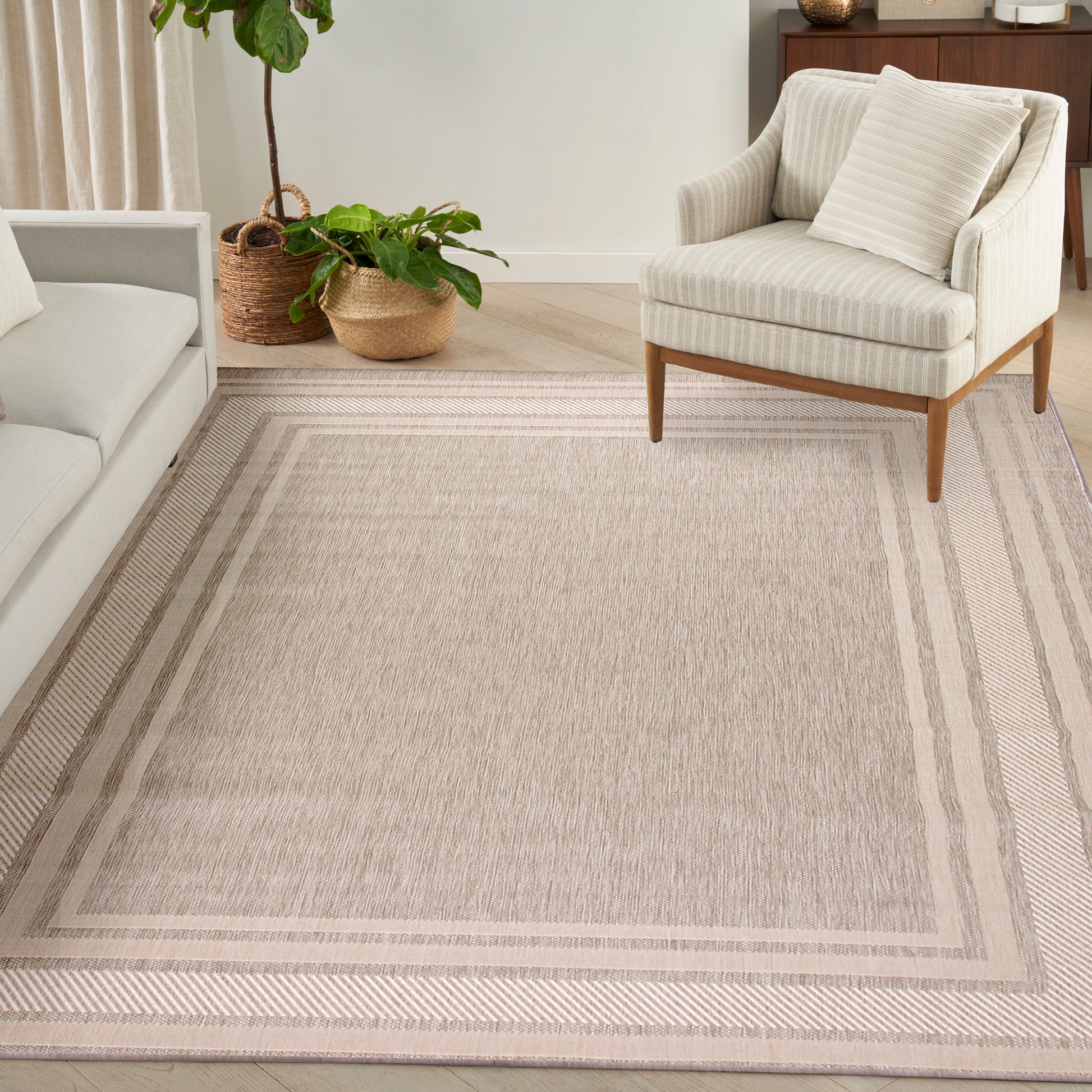 Beige textured area rug with double border stripes in a neutral living room, paired with a cream armchair and potted plants.