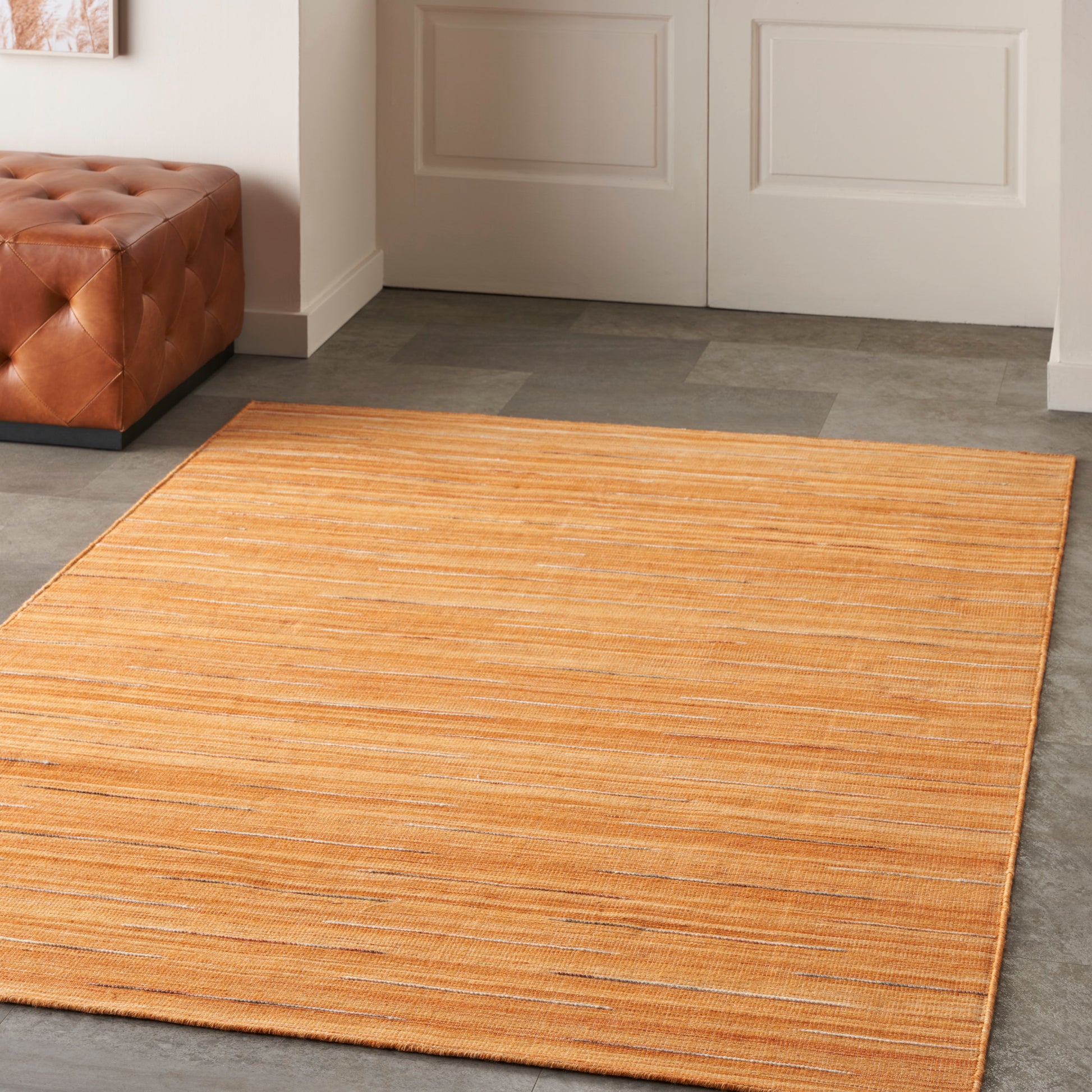 Orange flat-weave rug with subtle stripes on gray tile floor, beside a brown tufted leather bench and white paneled doors.