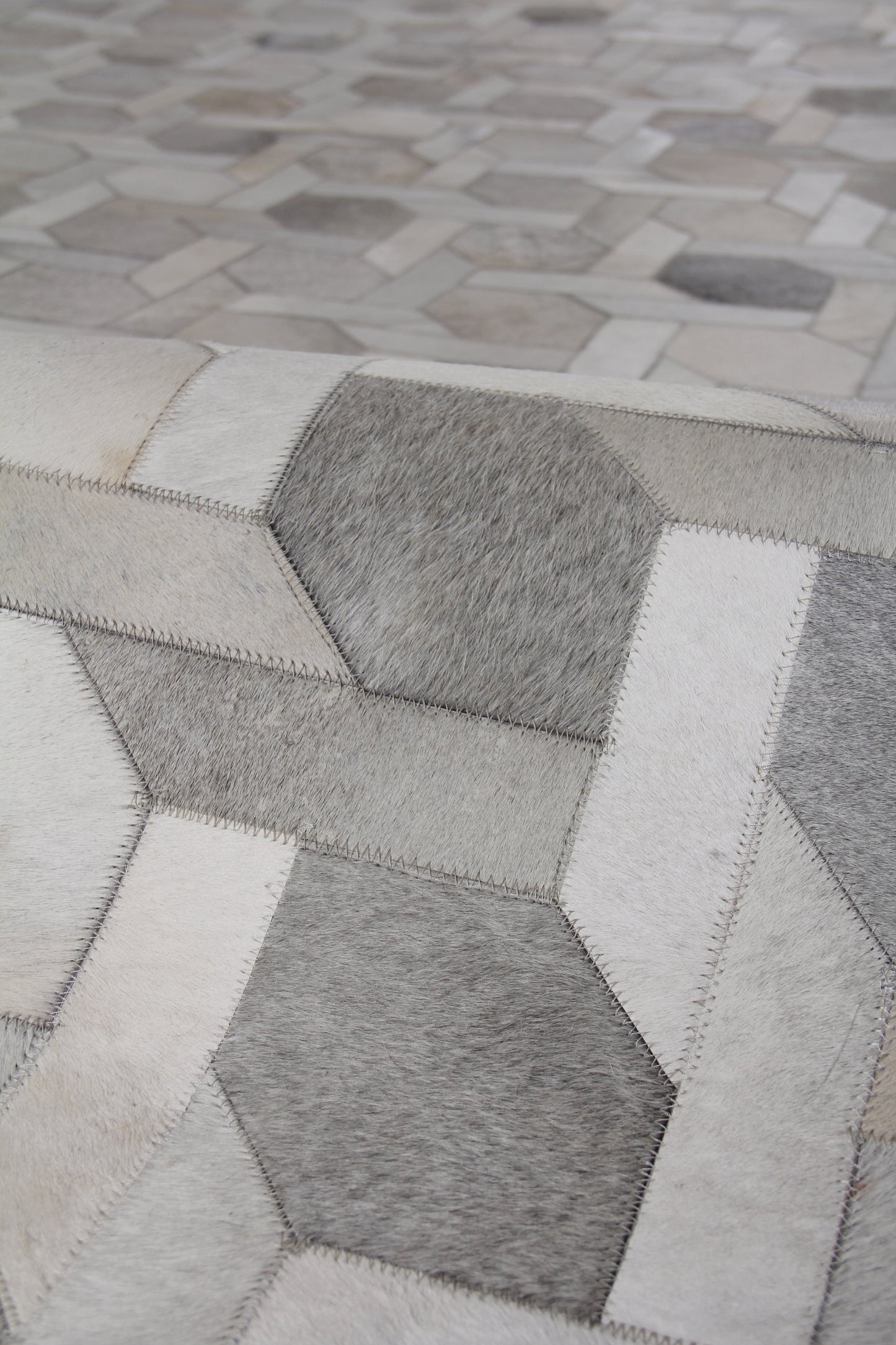 Close-up of a grayscale hexagon patchwork rug made from natural hide, hand-stitched, with light, medium, and dark gray patches and visible stitching.