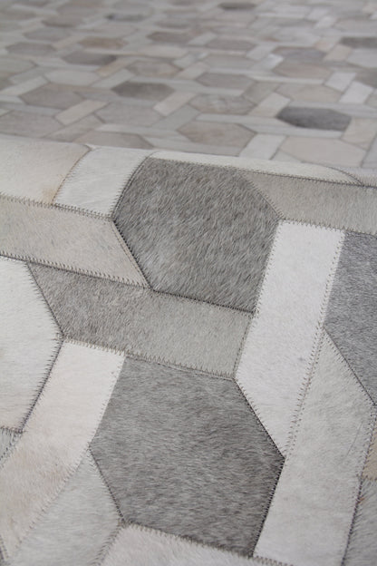 Close-up of a grayscale hexagon patchwork rug made from natural hide, hand-stitched, with light, medium, and dark gray patches and visible stitching.