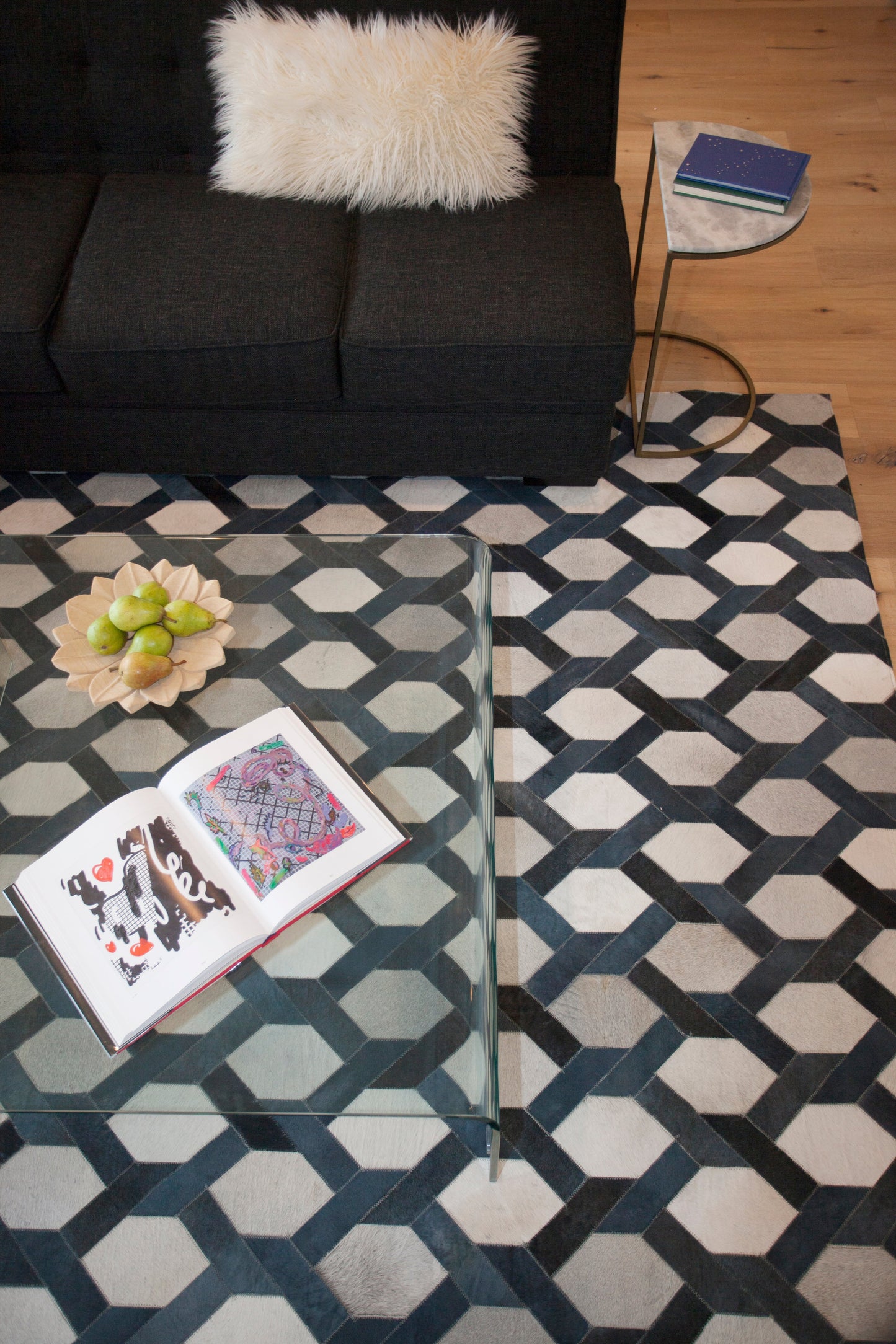 Dark gray sofa with fluffy white pillow, glass coffee table on a geometric navy rug, open art book and pear bowl.
