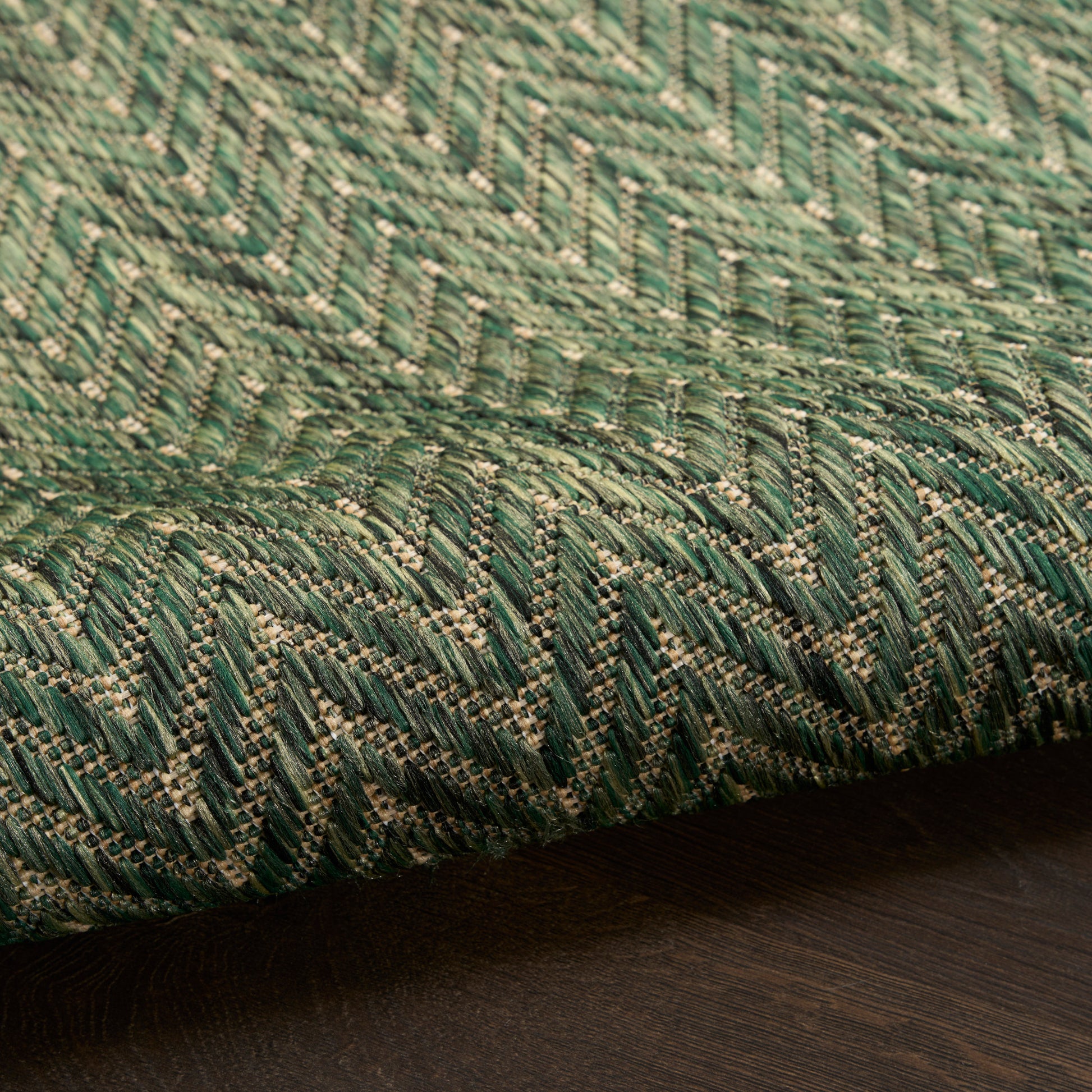 Close-up of green & beige chevron-patterned woven rug with textured yarns, edge curling on dark wood floor.