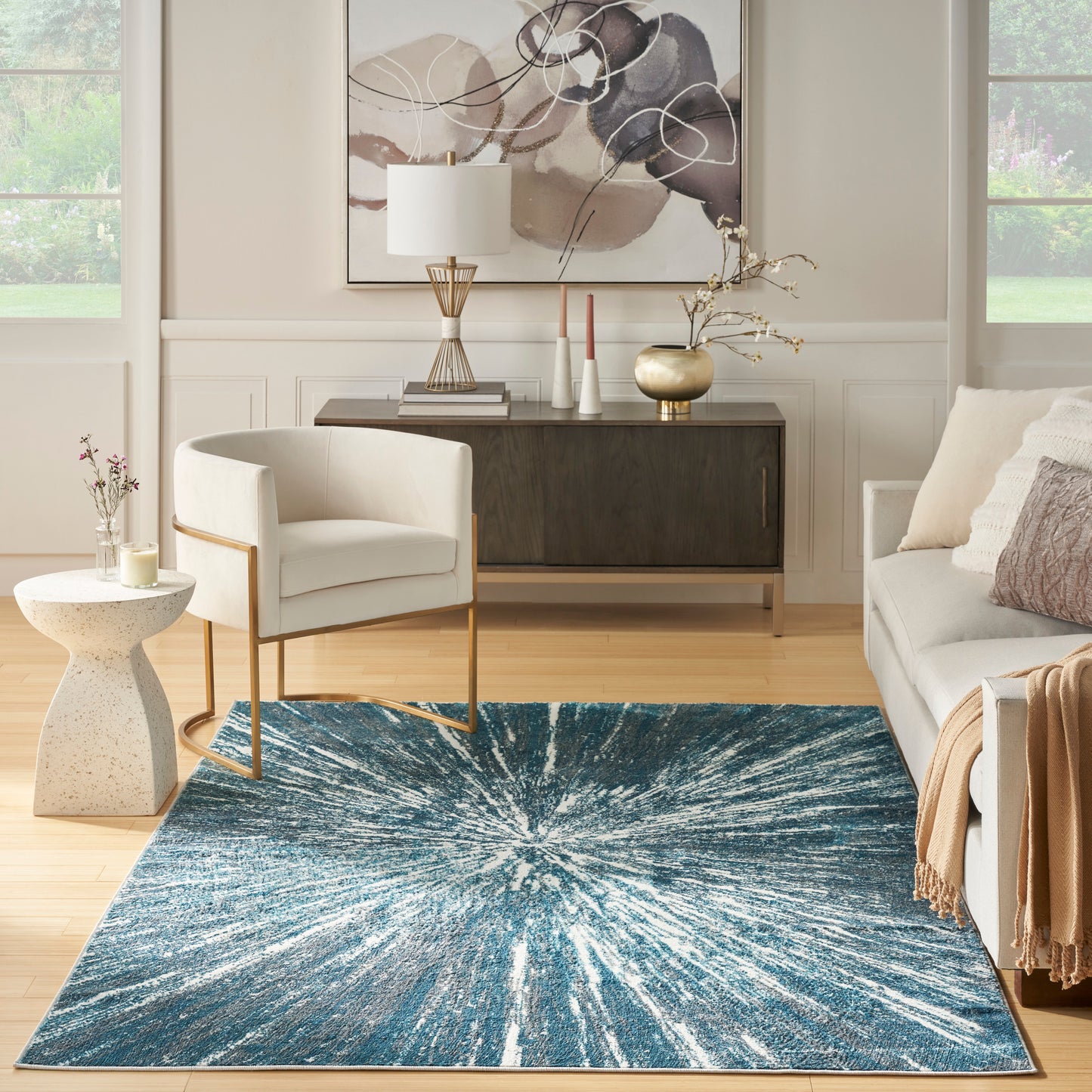 Modern living room with blue starburst rug, white armchair with gold legs, beige sofa, dark console, and abstract art.