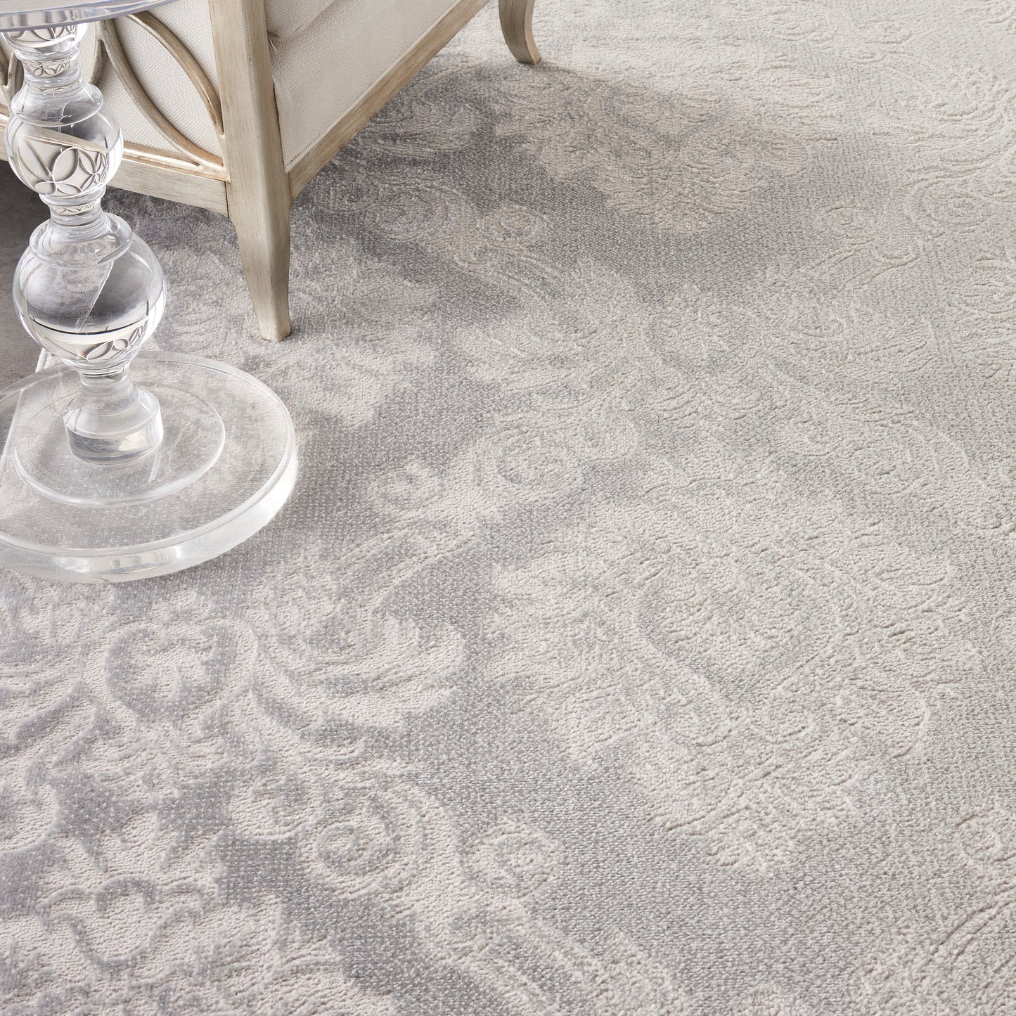 Light gray damask-pattern rug with floral motifs; crystal lamp base and beige furniture leg visible.