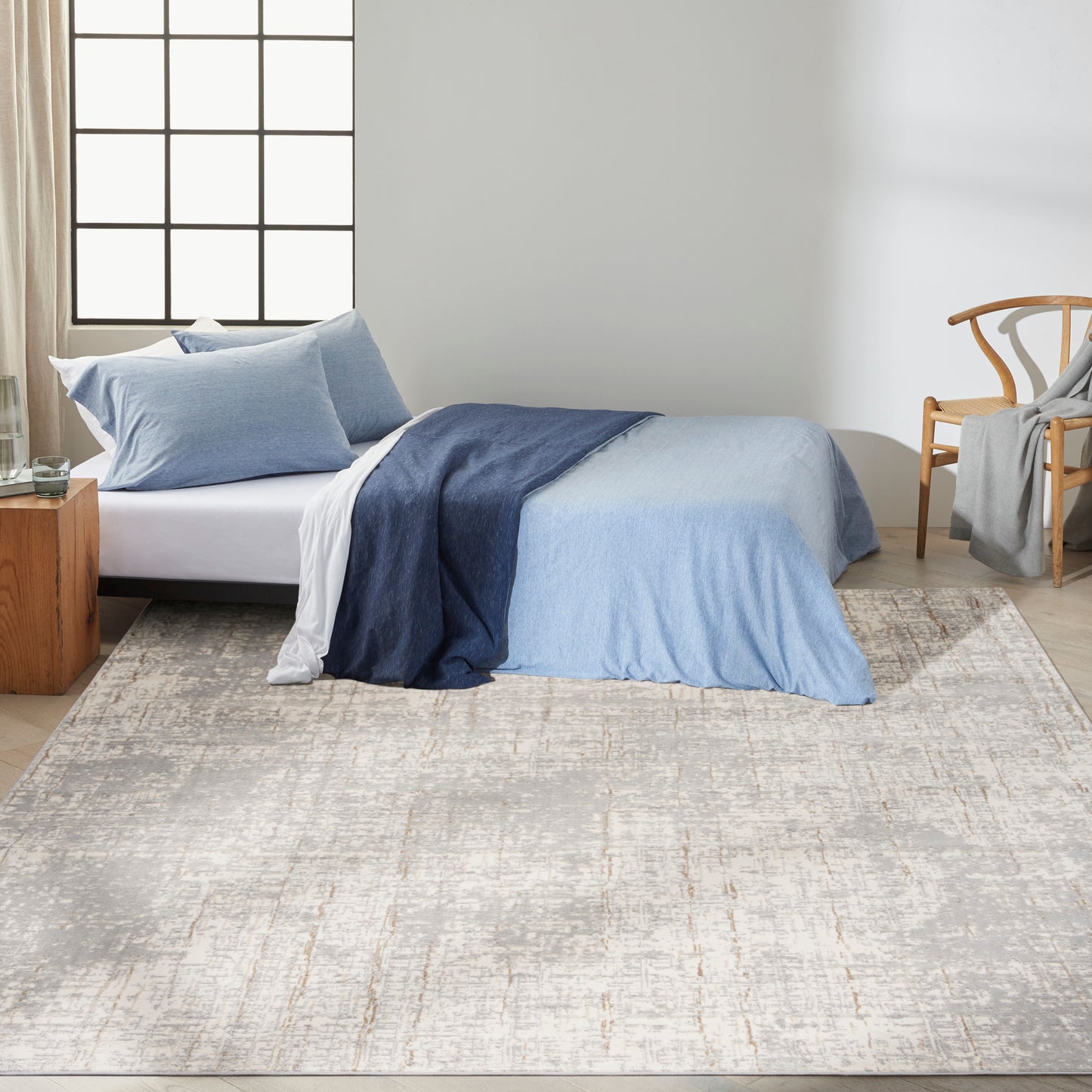 Bedroom scene with a beige patterned rug on the floor, a bed dressed in a blue gradient duvet cover with light blue pillows, white sheets, plus a wooden nightstand and a woven chair.