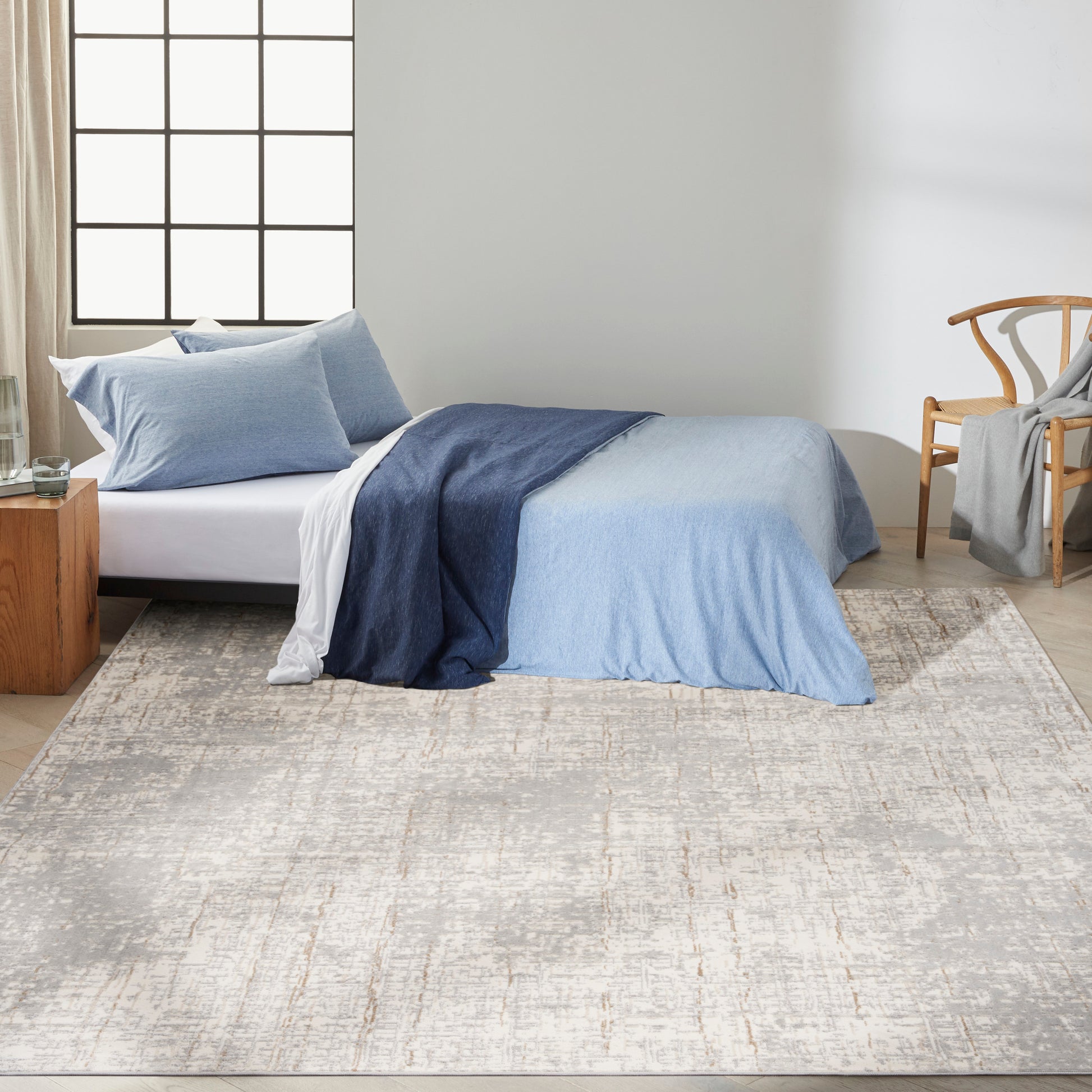 Bedroom scene with a beige patterned rug on the floor, a bed dressed in a blue gradient duvet cover with light blue pillows, white sheets, plus a wooden nightstand and a woven chair.