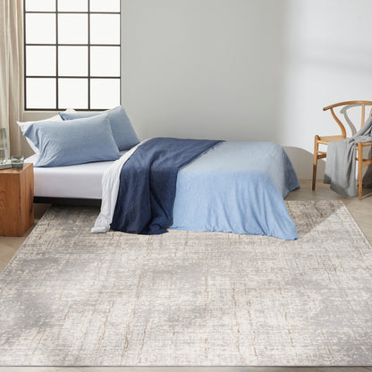 Bedroom scene with a beige patterned rug on the floor, a bed dressed in a blue gradient duvet cover with light blue pillows, white sheets, plus a wooden nightstand and a woven chair.