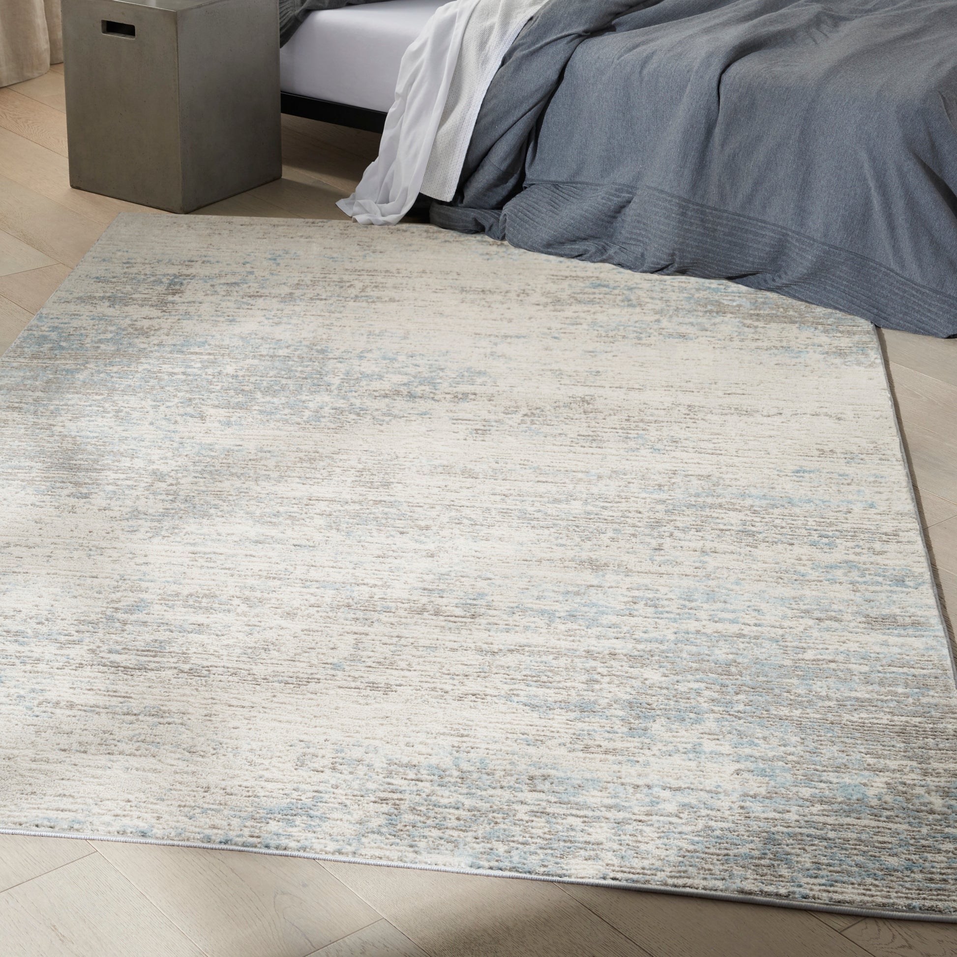 Rectangular flat-weave area rug with beige, gray & pale blue mottling on light wood floor beside a bed.