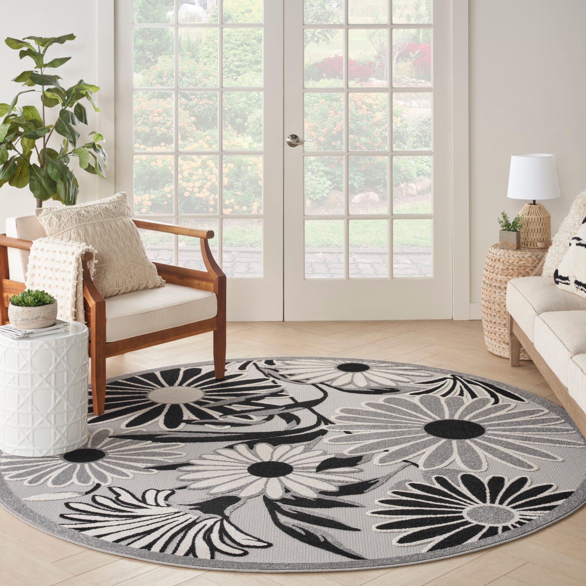 Round area rug with large black and white floral pattern on gray, placed in a bright living room beside chair and sofa.