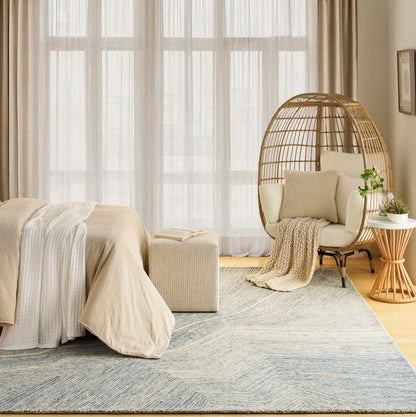 Beige-toned bedroom with large windows, linen bed, woven rattan egg chair, cozy throws, blue patterned rug, plants.