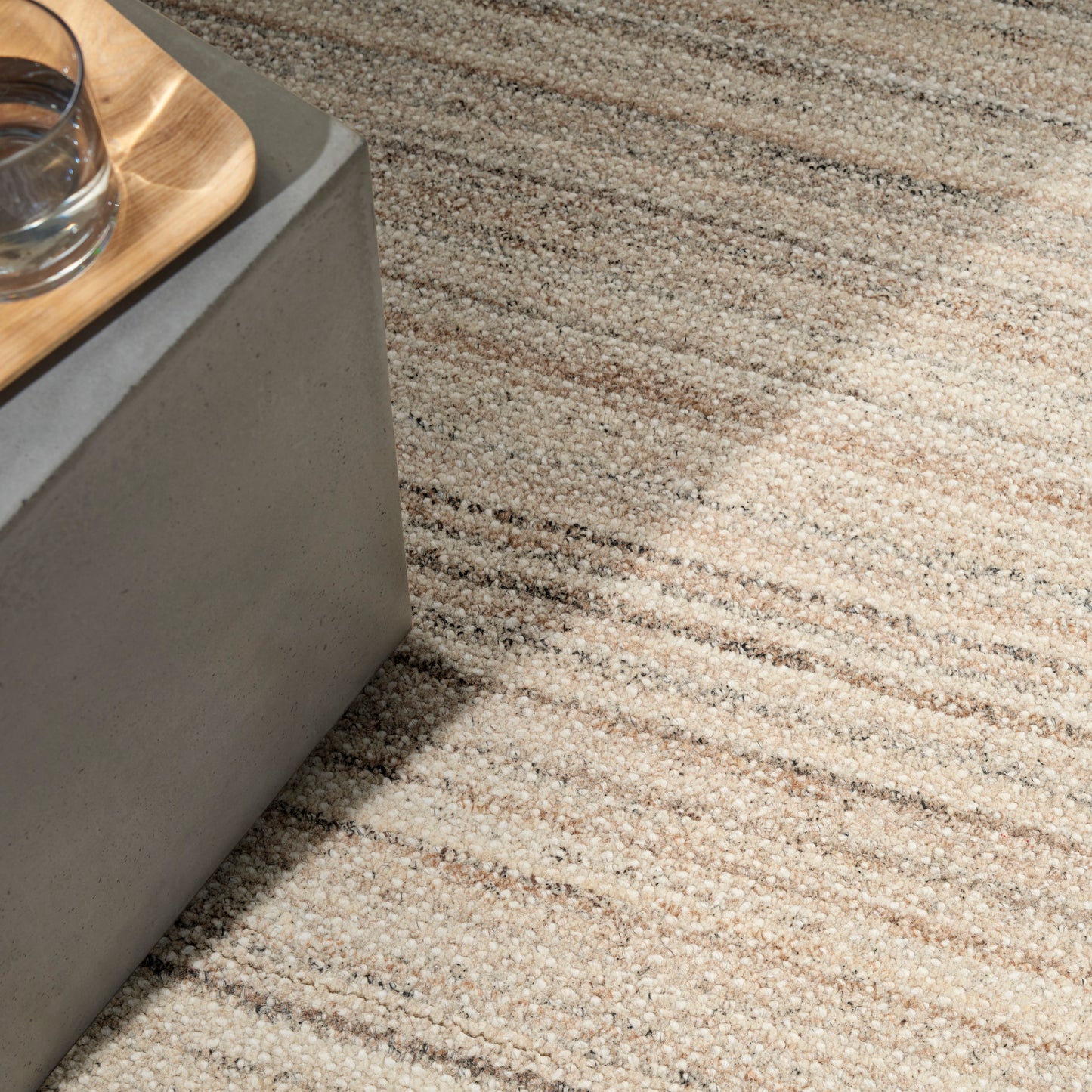 Beige striped textured rug with loop pile; gray concrete coffee table and wooden tray with glass.