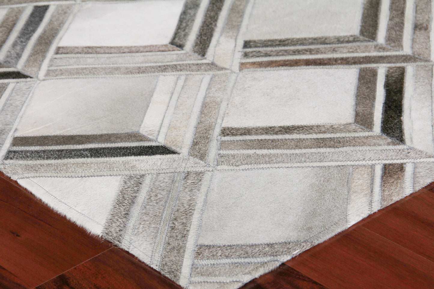 Natural hide indoor hand-stitched rug on wood floor; light gray tones with a subtle irregular pattern and stitched edge.