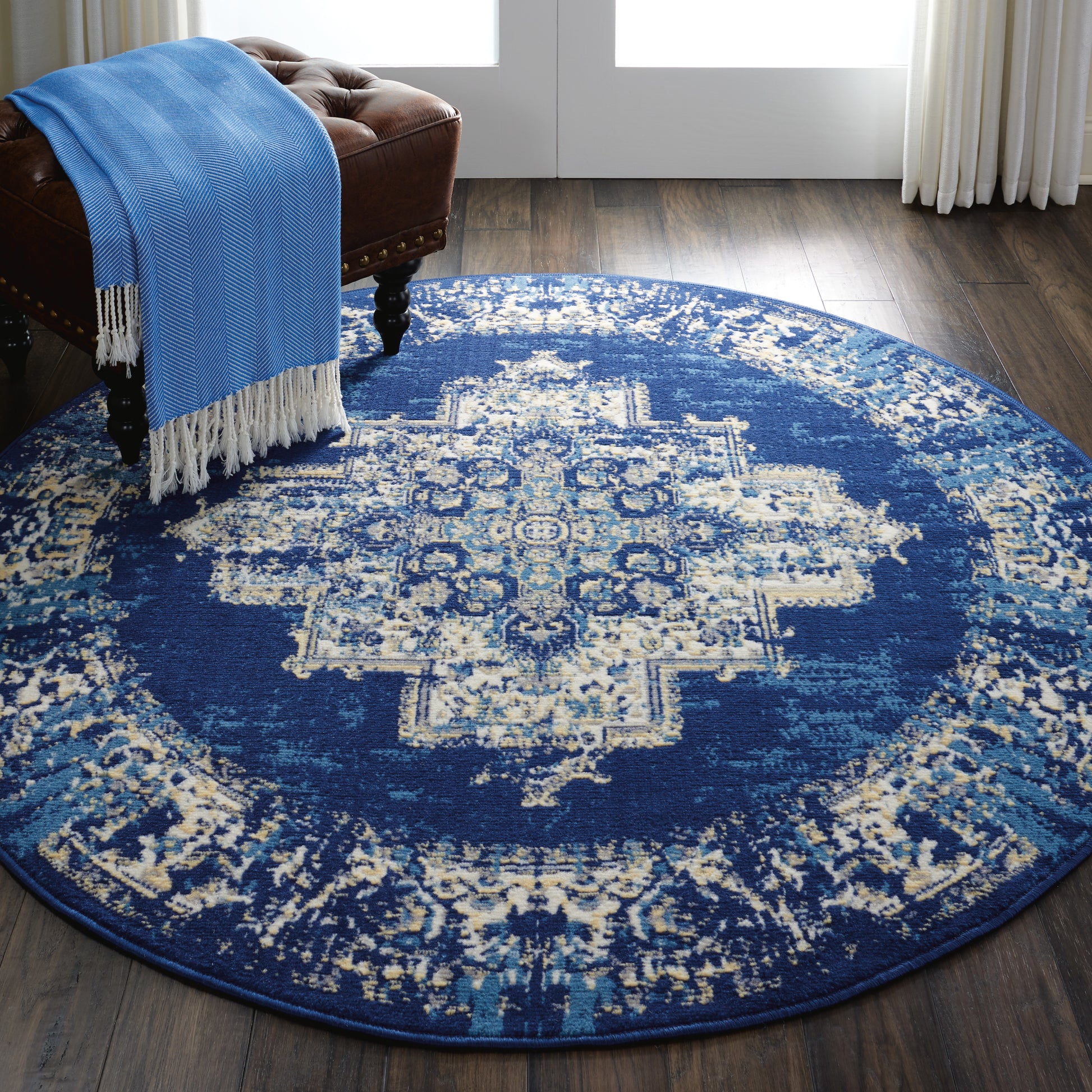 Round navy blue oriental rug with ivory distressed medallion pattern, paired with a tufted brown leather ottoman and blue throw.