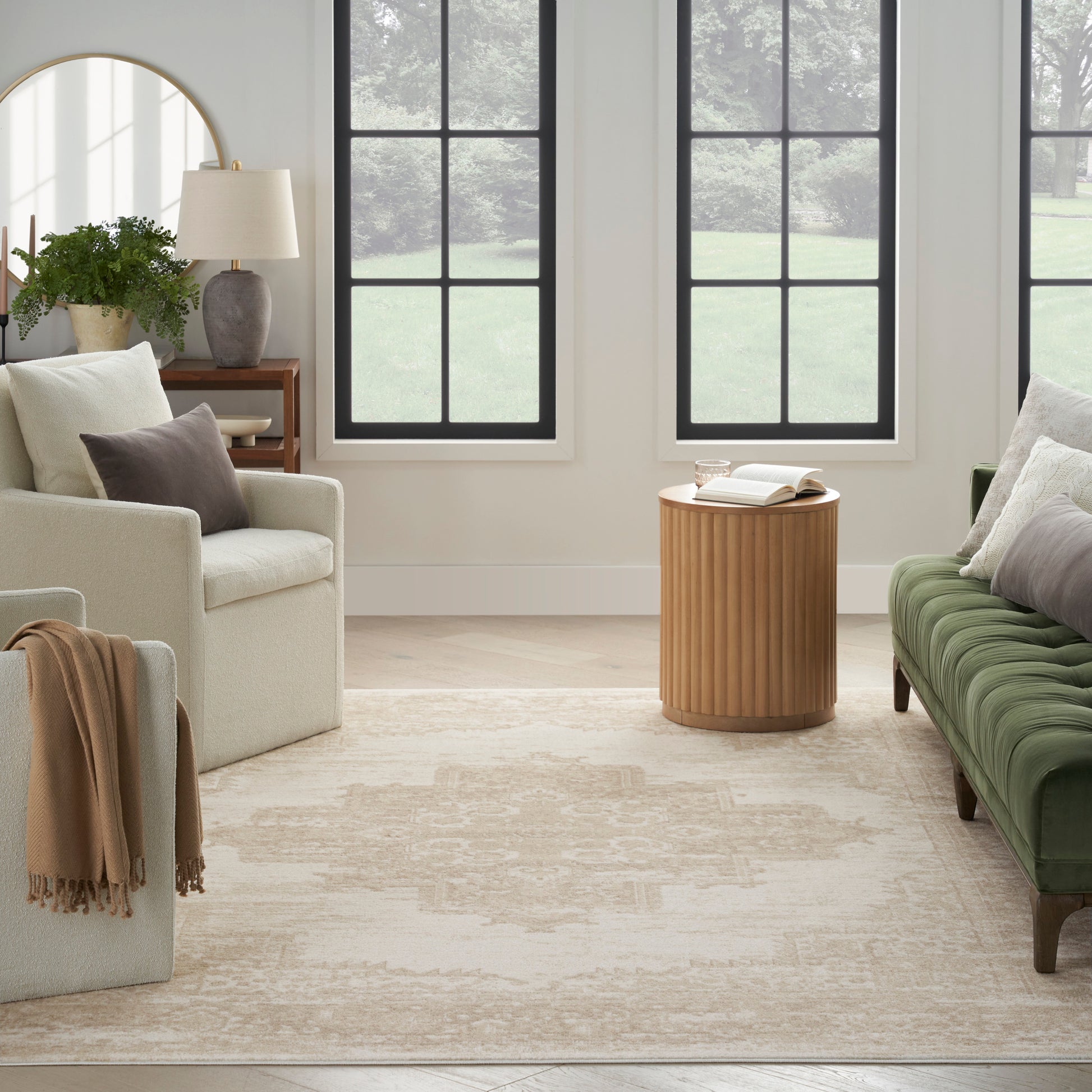 Cozy neutral living room with cream chairs, green velvet sofa, round ribbed wood table, beige rug, black-framed windows.