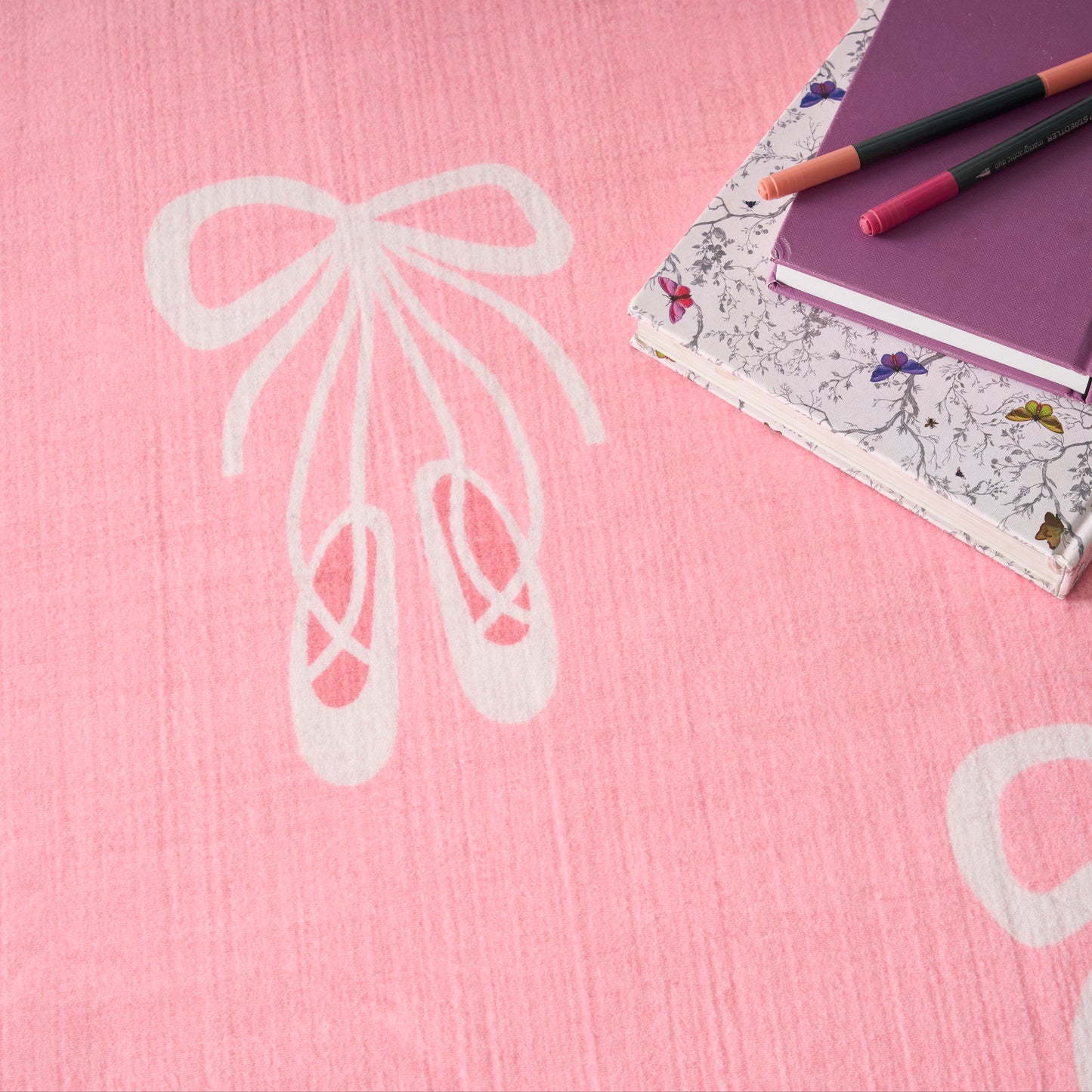 Pink textile rug with large white bow design, beside floral-edge notebooks and a purple notebook with pens.